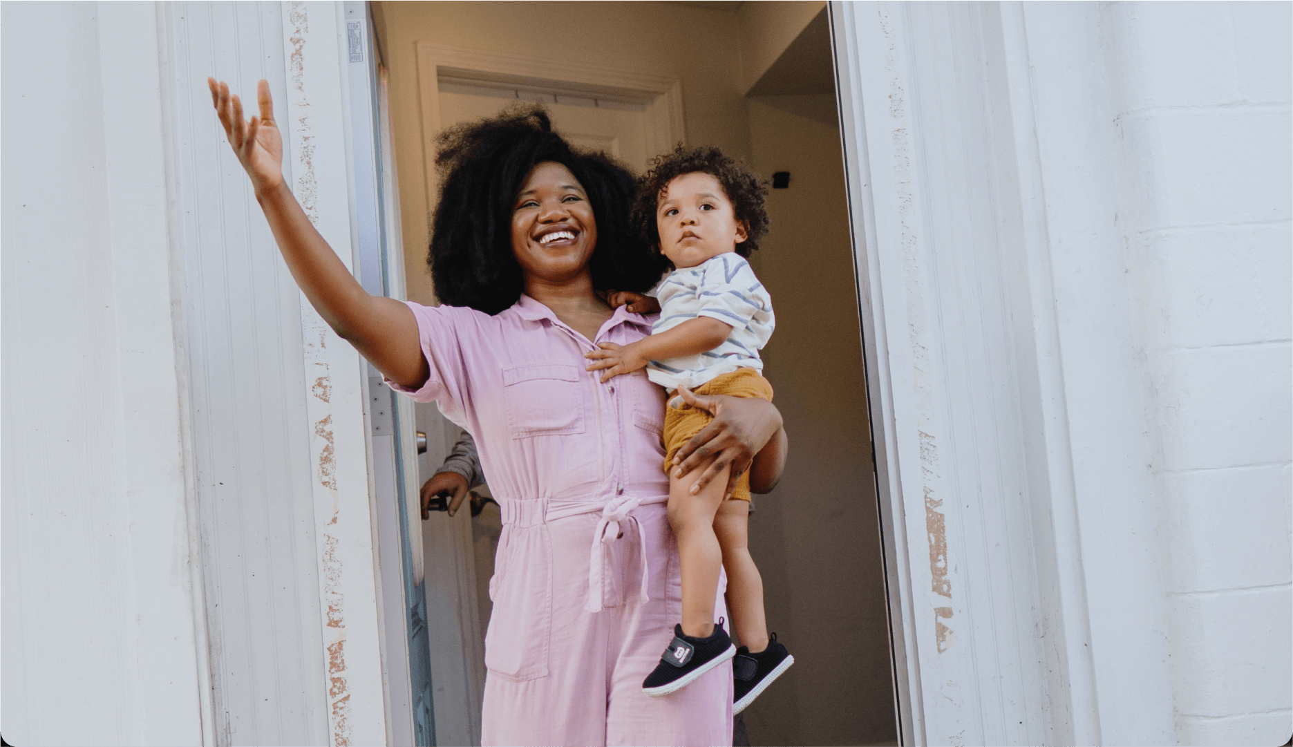 A smiling woman in a pink outfit holding a young child waves cheerfully while standing in the doorway of a white house.