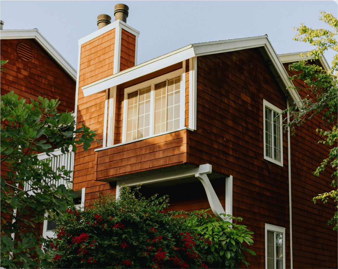 A two-story house with warm reddish-brown wooden shingles and white trim, surrounded by greenery and flowering bushes.