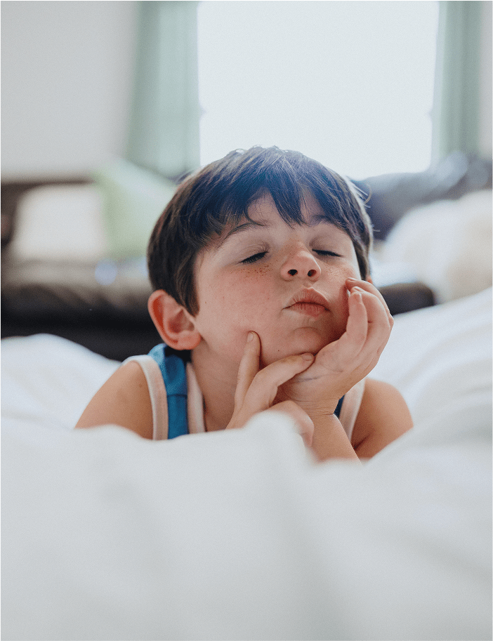 A young boy with short dark hair lies on a bed, eyes closed and hands resting under his chin, looking peaceful and thoughtful.