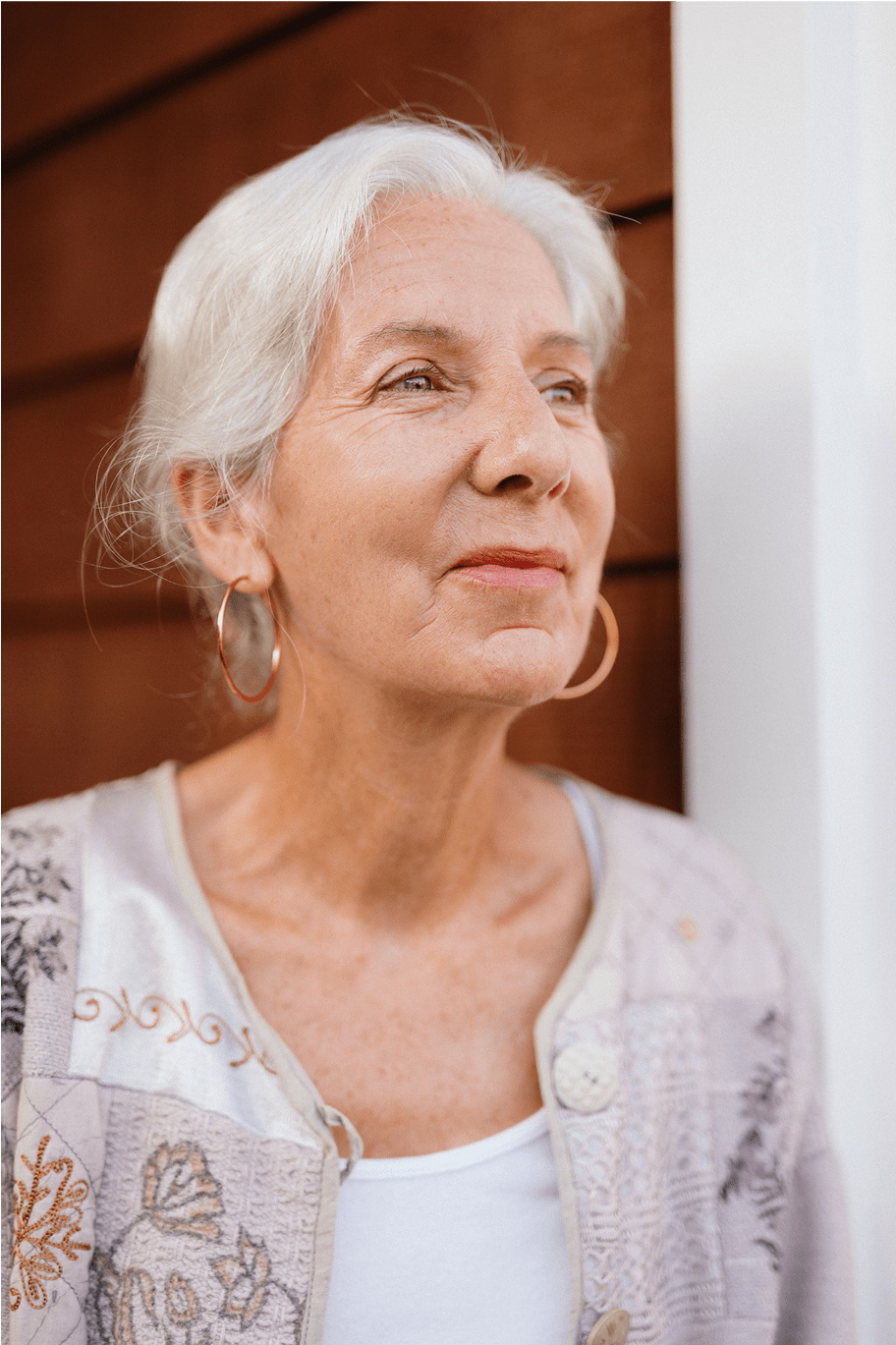 A close-up of an older woman with white hair and hoop earrings standing outside against a wooden wall, gazing upward with a serene expression.