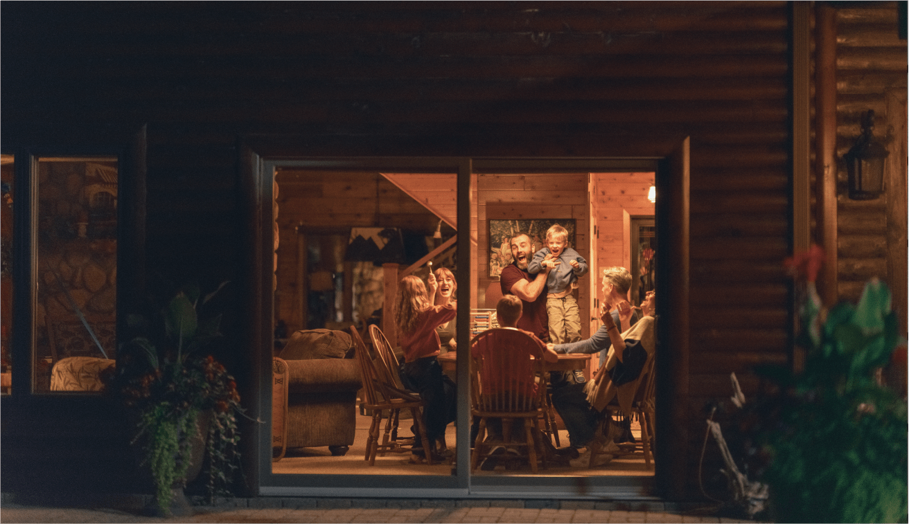 A warm evening scene viewed through large windows, showing a group of people gathered around a wooden dining table inside a cozy log cabin, laughing and enjoying each other’s company.