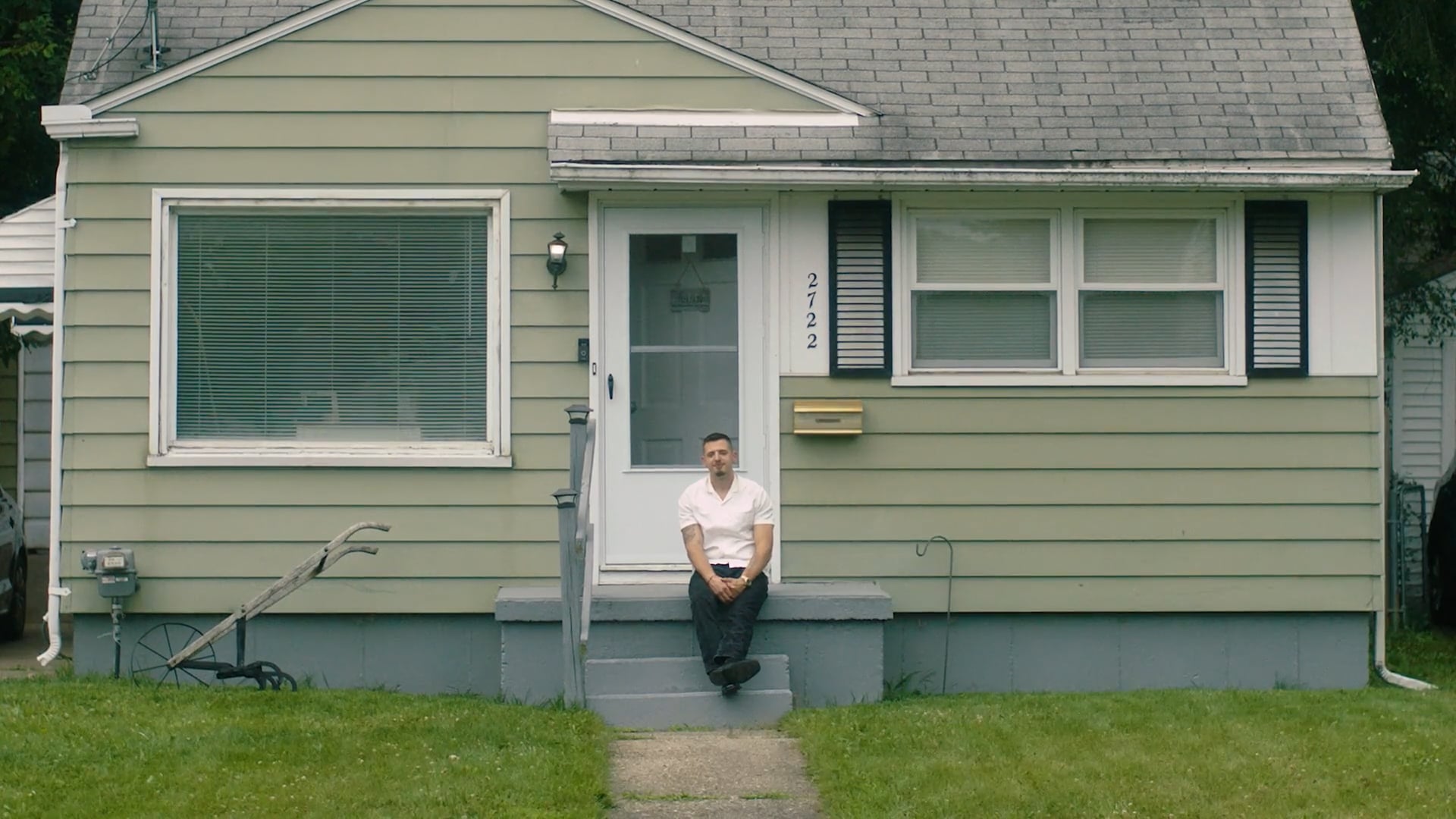A man in a white shirt sits alone on the front steps of a small green house with white trim. He looks thoughtful or contemplative, framed by the quiet, suburban setting.
