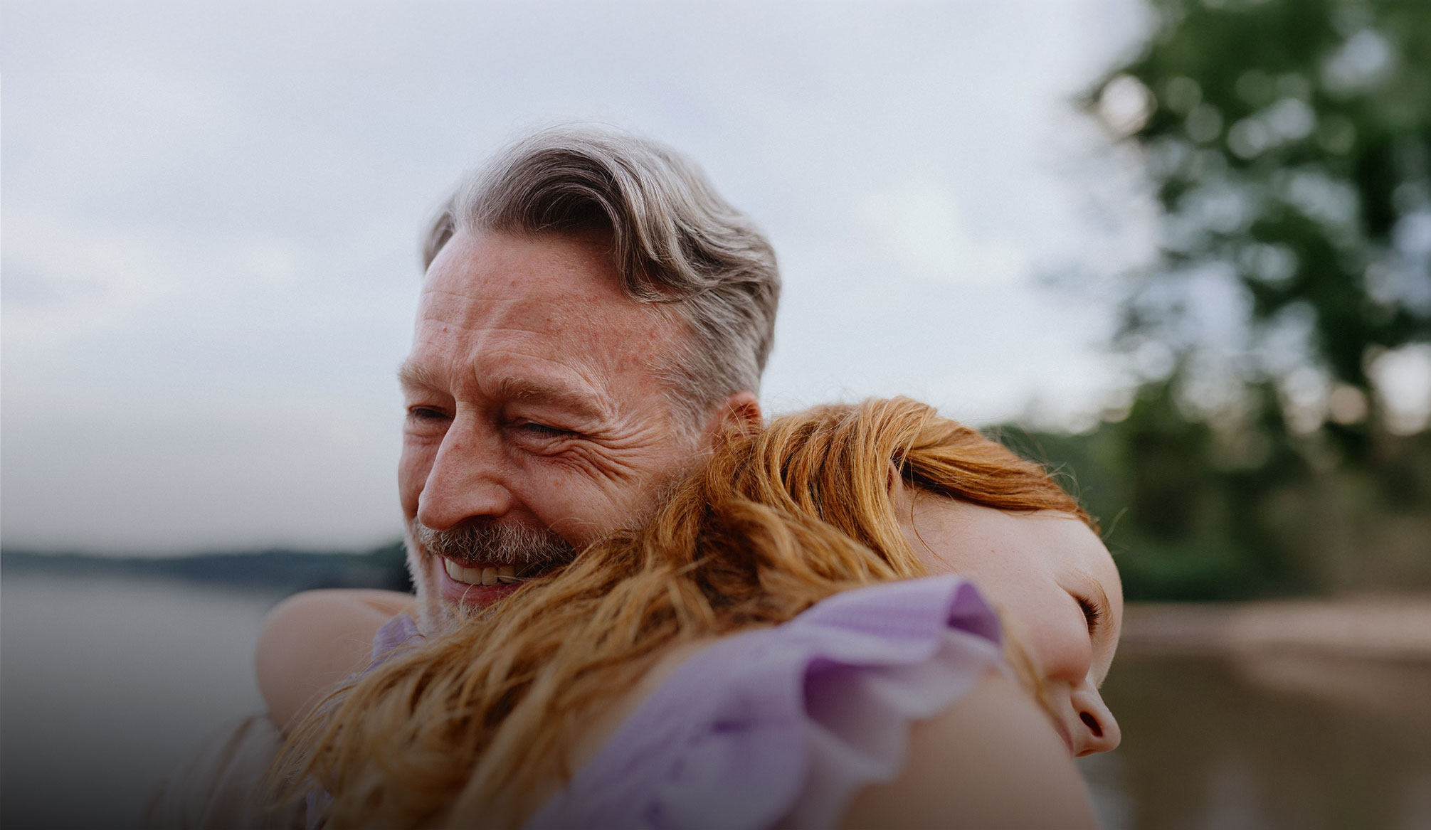 An older man with gray hair smiles warmly while hugging a young girl with red hair outdoors near a lake.