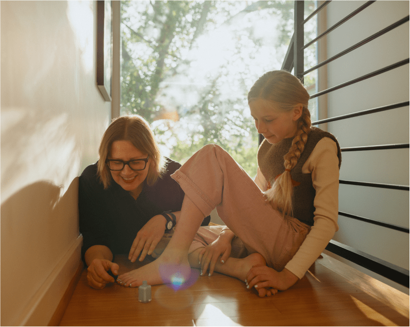 A smiling woman and a young girl with a braid sit on the floor by a sunny window, as the woman paints the girl’s toenails, both enjoying the quiet moment together.