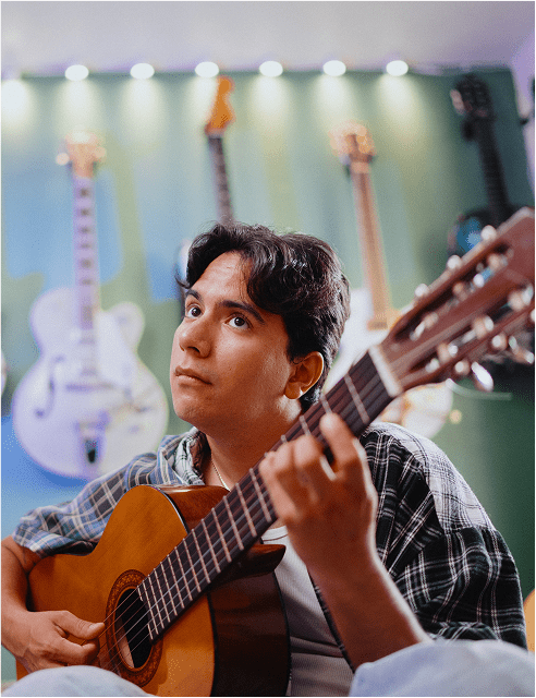 A young man strums an acoustic guitar while seated in a room with several guitars hanging on the wall behind him, looking thoughtful as he plays.