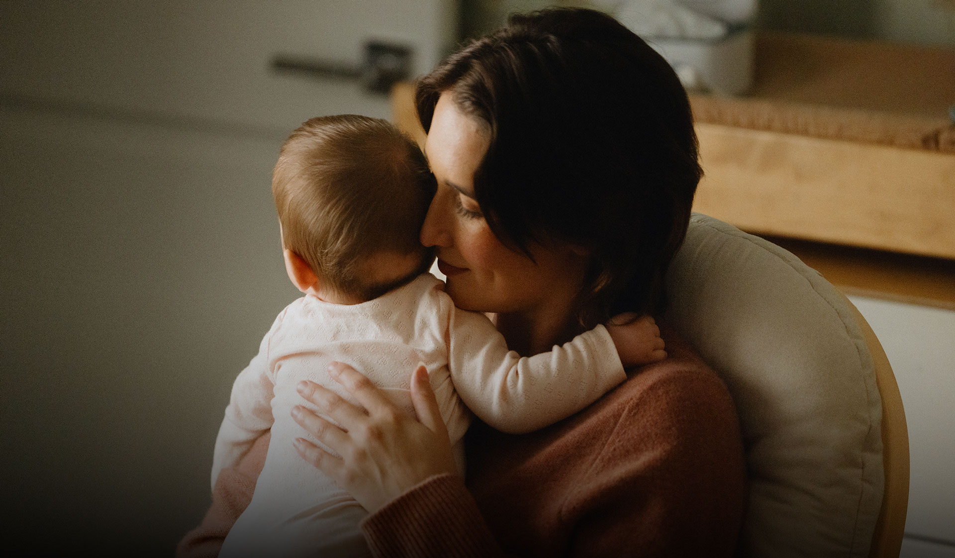 Close-up of a woman lovingly holding a baby against her shoulder, softly smiling in a cozy indoor setting.