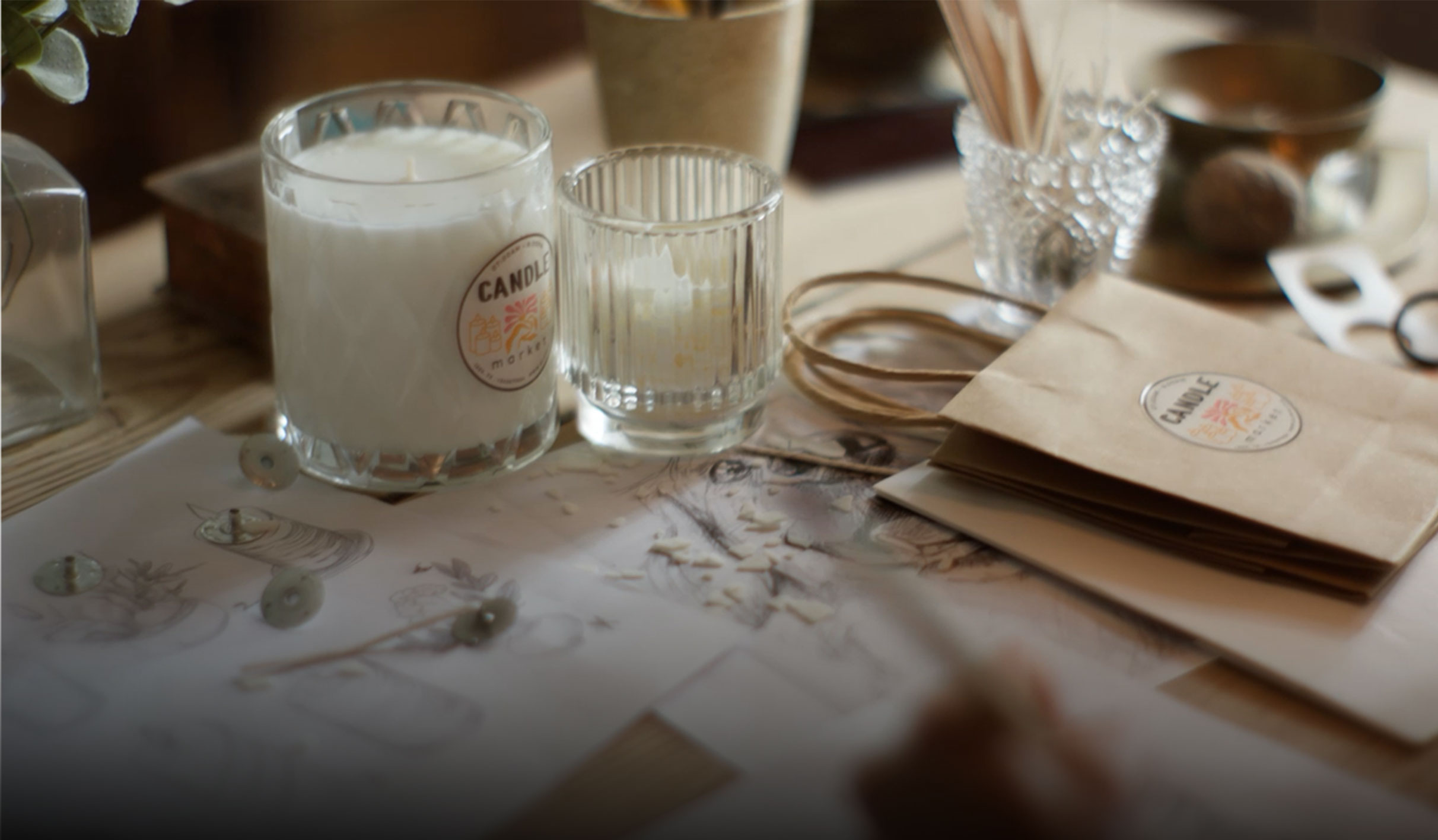 A tabletop workspace with sketches, candles, jars, scissors, and craft supplies, suggesting candle-making.