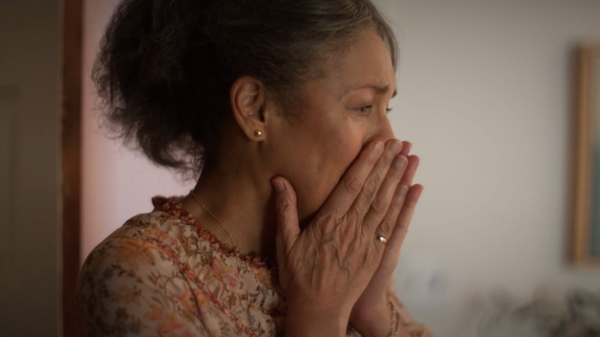 A woman covering her mouth with her hands in emotional surprise, wearing a floral blouse and small gold earrings.