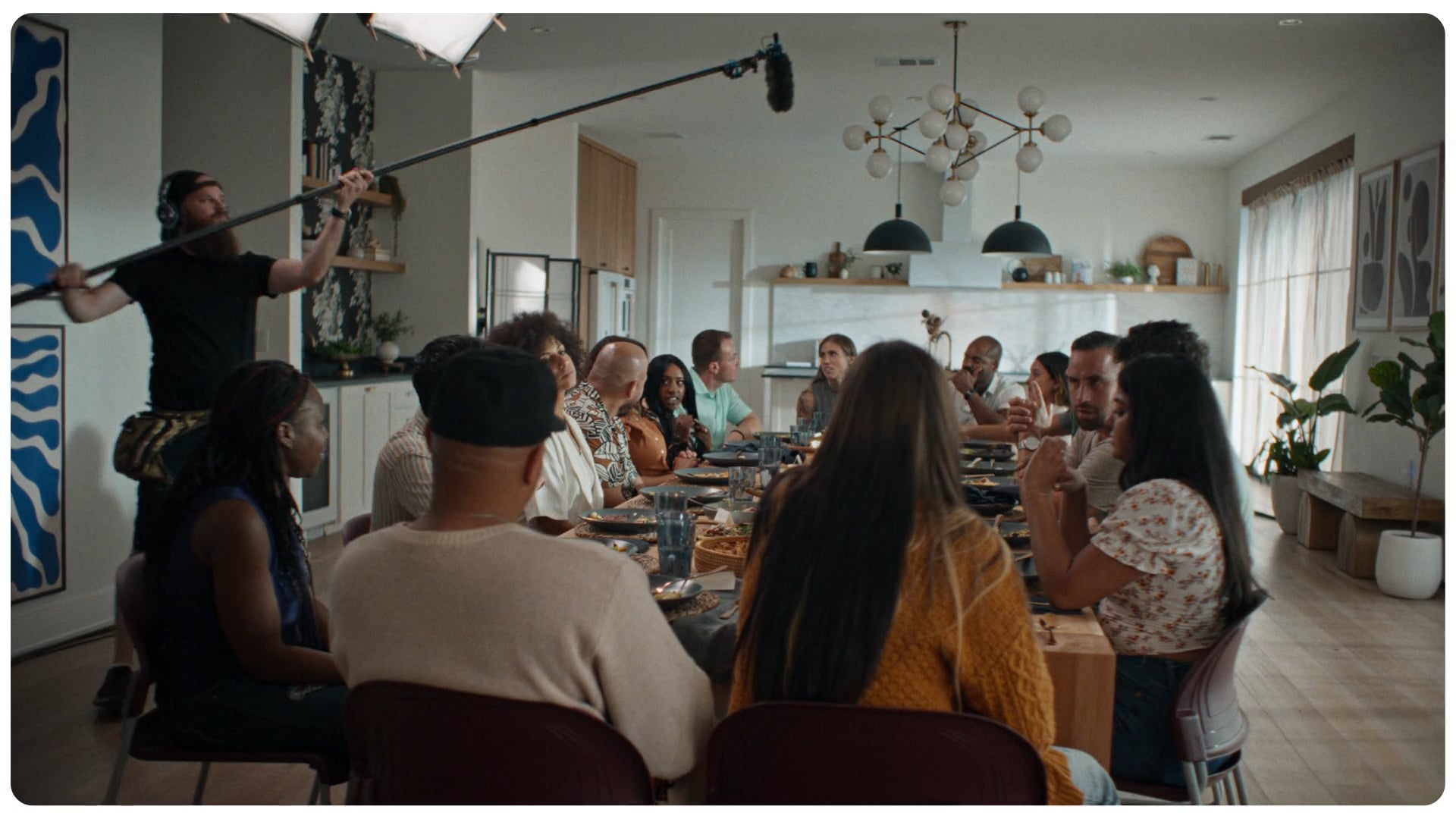 A behind-the-scenes shot of a diverse group gathered around the same dining table. A boom microphone operator and lighting equipment are visible, showing a film production in progress.
