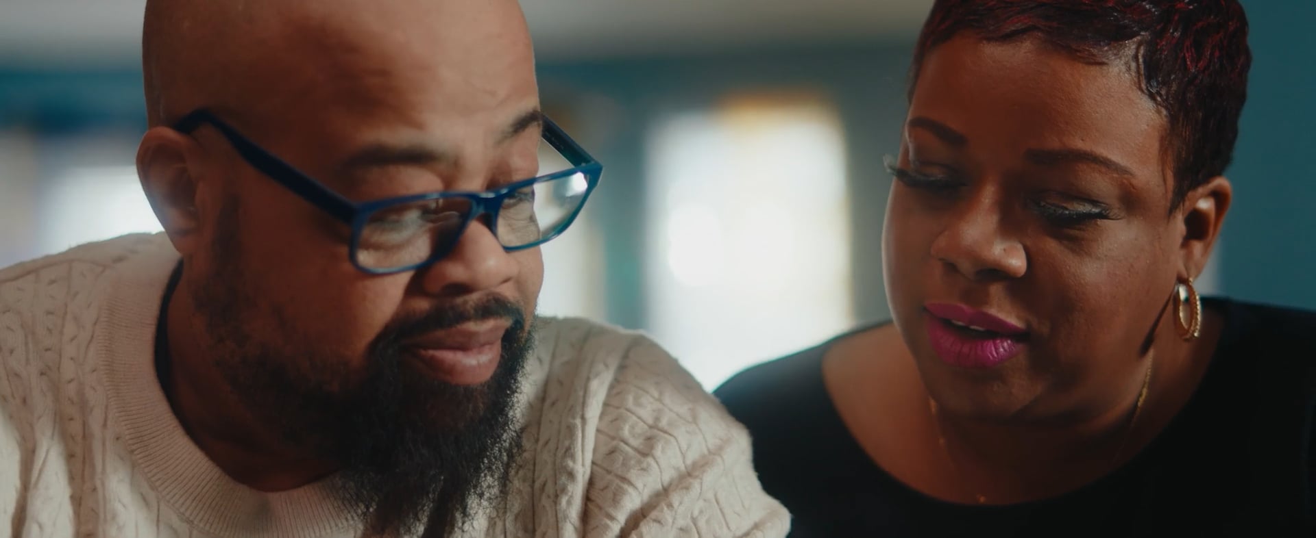 A close-up of a man with glasses and a beard wearing a cream sweater, sitting beside a woman with short hair, gold hoop earrings, and pink lipstick. They appear to be having a thoughtful conversation.