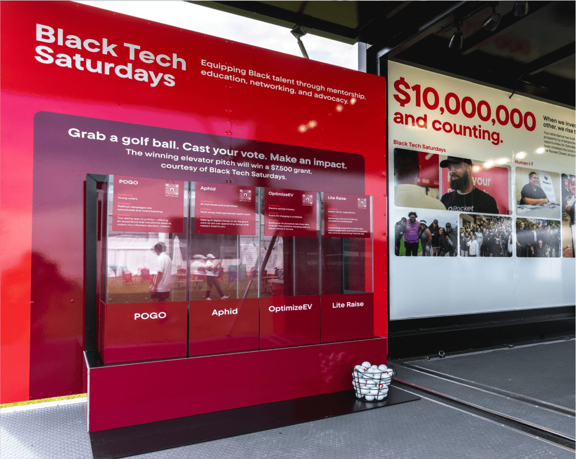 A red and white exhibit display for Black Tech Saturdays featuring startup pitch information and signs promoting mentorship, education, and advocacy, with golf balls in a basket at the bottom.