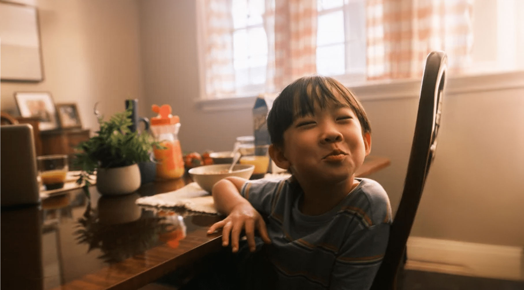 A young boy sits at a dining table with breakfast items, smiling mischievously while sunlight streams through curtained windows behind him.