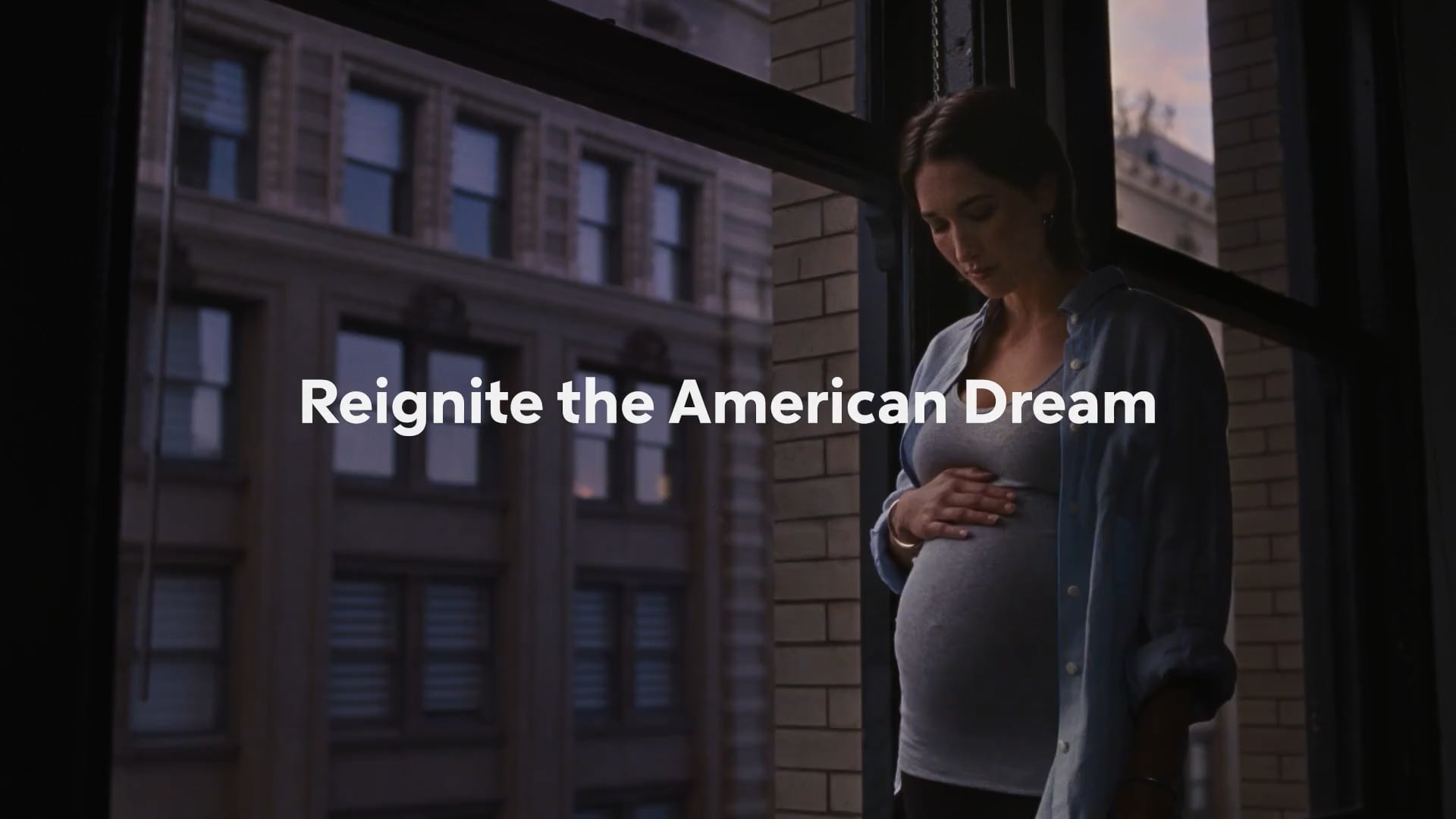 A pregnant woman stands by a large window in a city apartment, gently resting her hand on her belly with the words “Reignite the American Dream” overlaid.