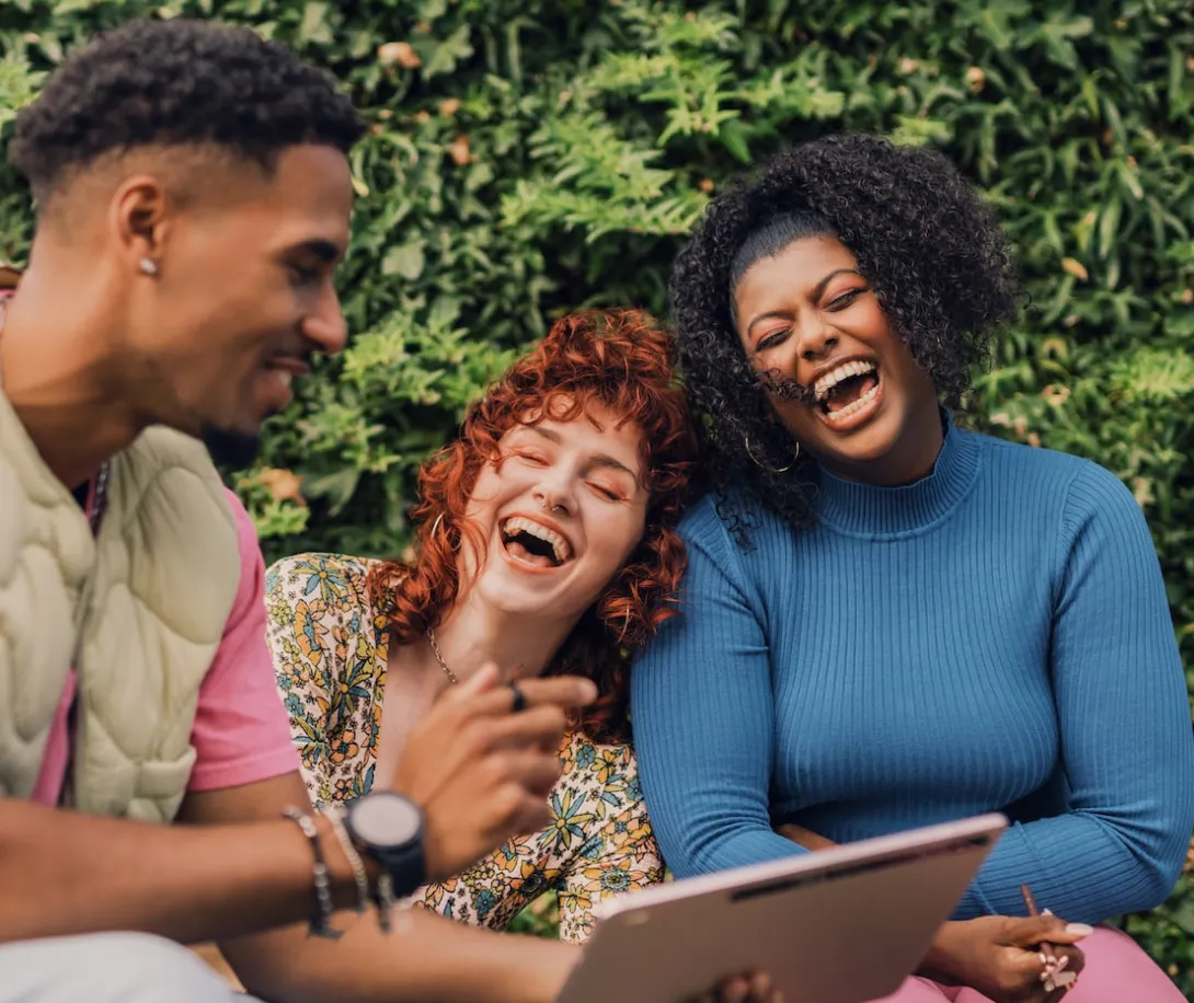 Three friends sitting outdoors laughing while looking at a tablet together.