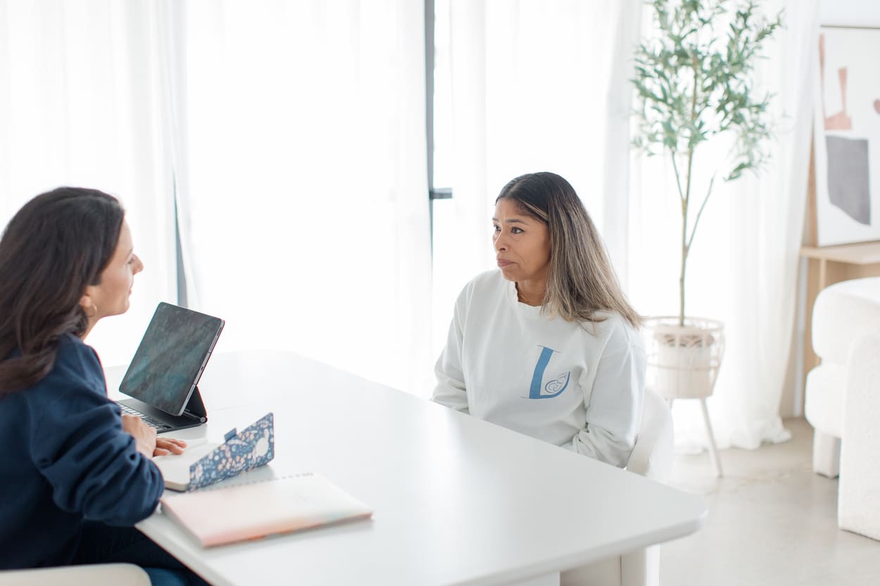 Two women seated across a white table in a bright room, one attentively listening while the other expresses concern.