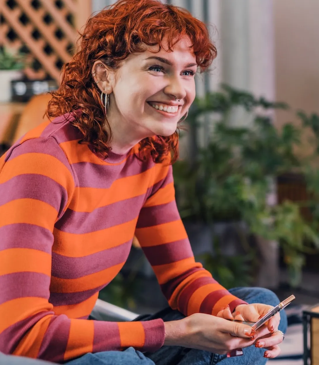 Smiling red-haired woman in orange and purple striped sweater sitting and holding a vape pen indoors.