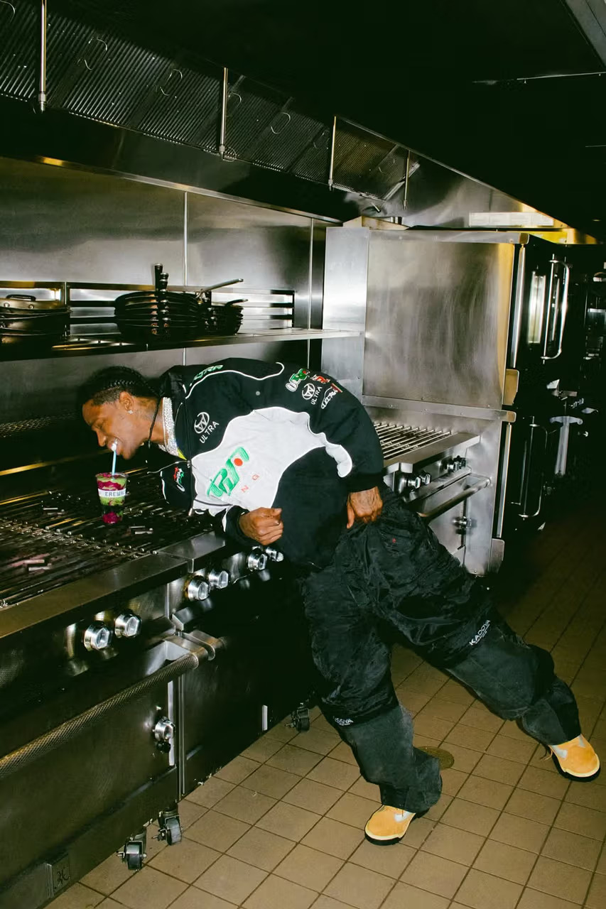 Travis Scott leaning over a commercial kitchen stove sipping an Erewhon Storm Smoothie, wearing a graphic racing jacket as part of the Cactus Jack x Erewhon campaign