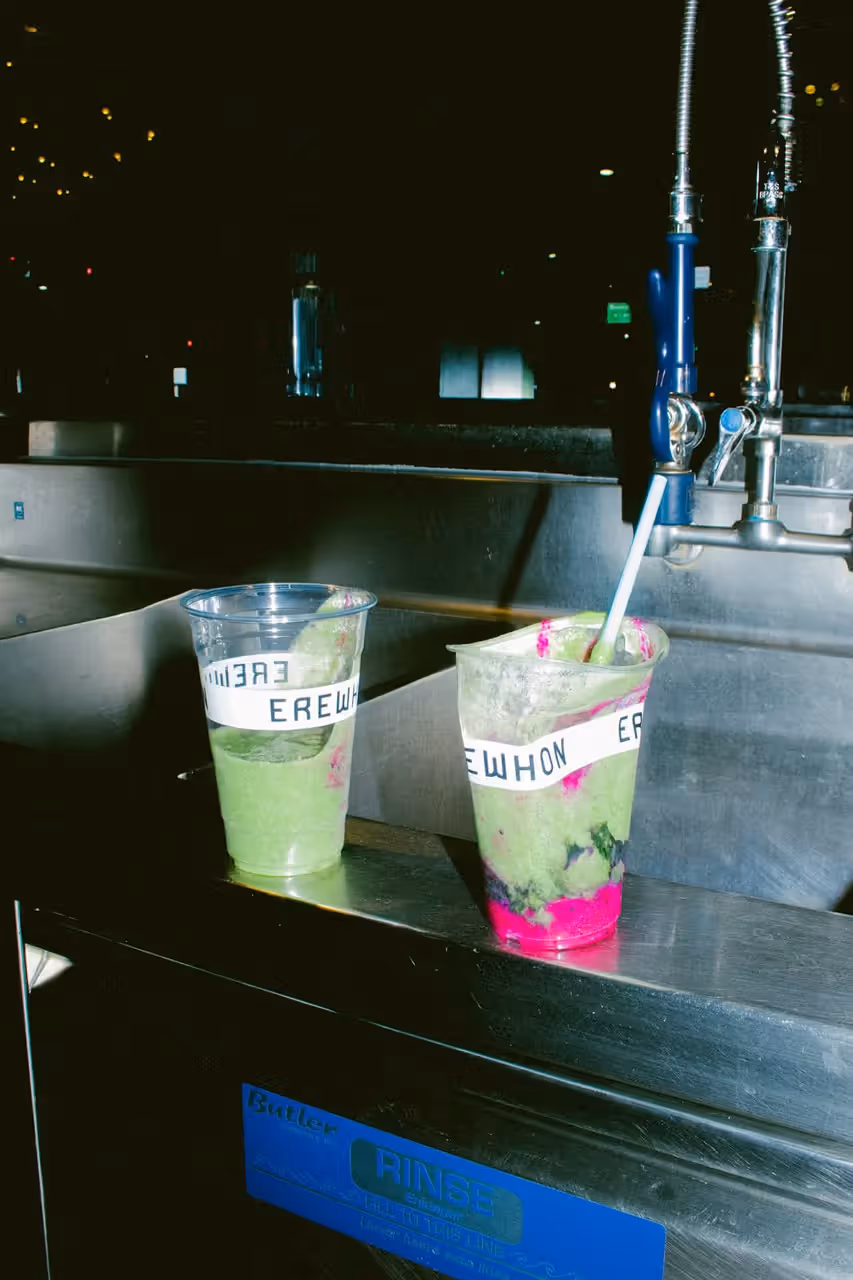 Two Erewhon branded cups containing the Travis Scott Storm Smoothie with green and pink layers resting on a stainless steel kitchen counter at night