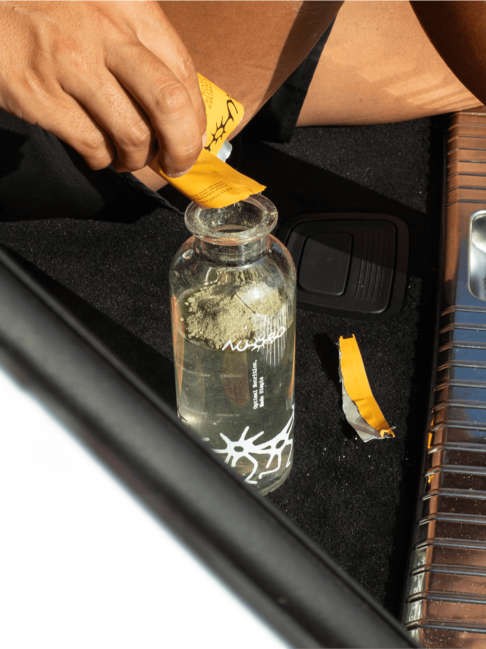 Lifestyle shot of a Nuoro supplement sachet being poured into a branded glass bottle, photographed in a vehicle setting