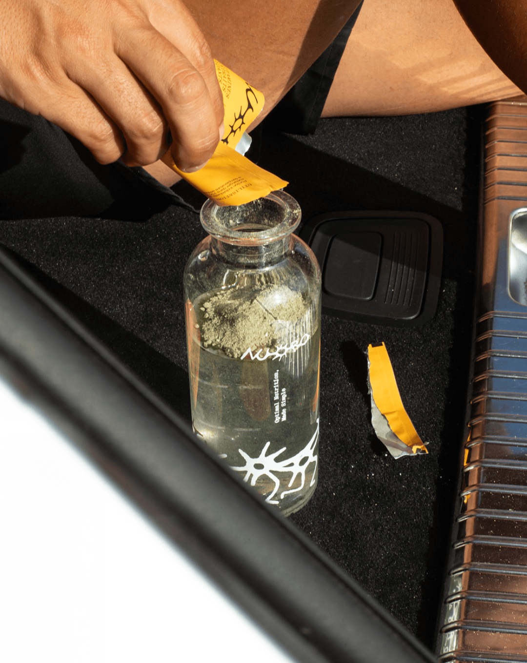 Lifestyle shot of a Nuoro supplement sachet being poured into a branded glass bottle, photographed in a vehicle setting