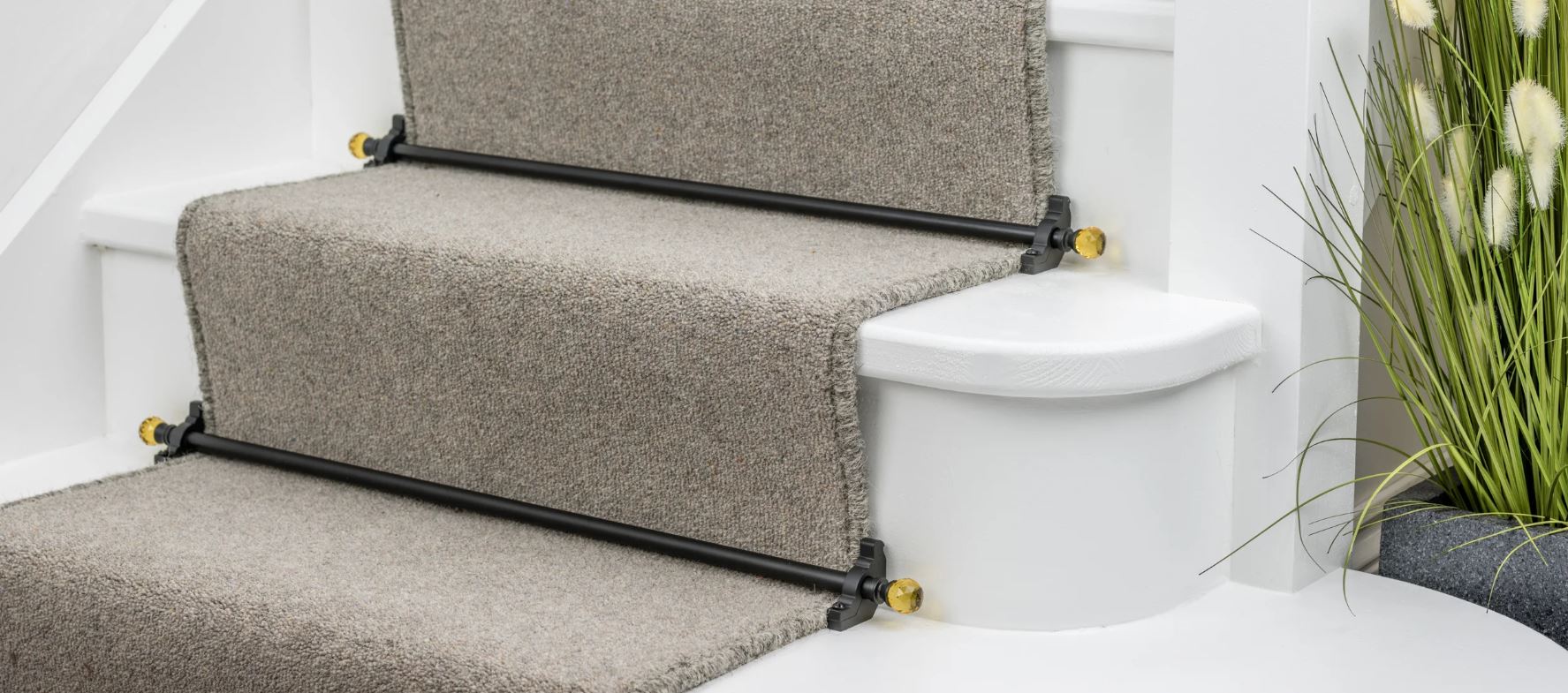 Close-up of beige carpet stair runner secured by black rods with decorative brass ends on white wooden stairs next to a plant.