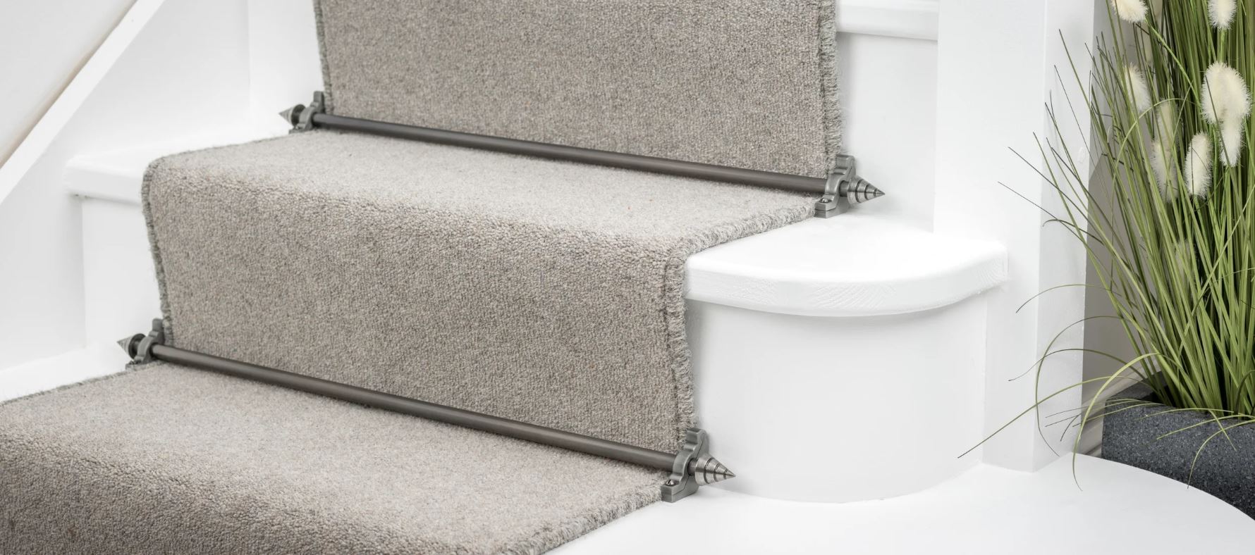 Close-up of beige carpet stair runners secured with metal stair rods on white wooden stairs, with decorative green plants beside.