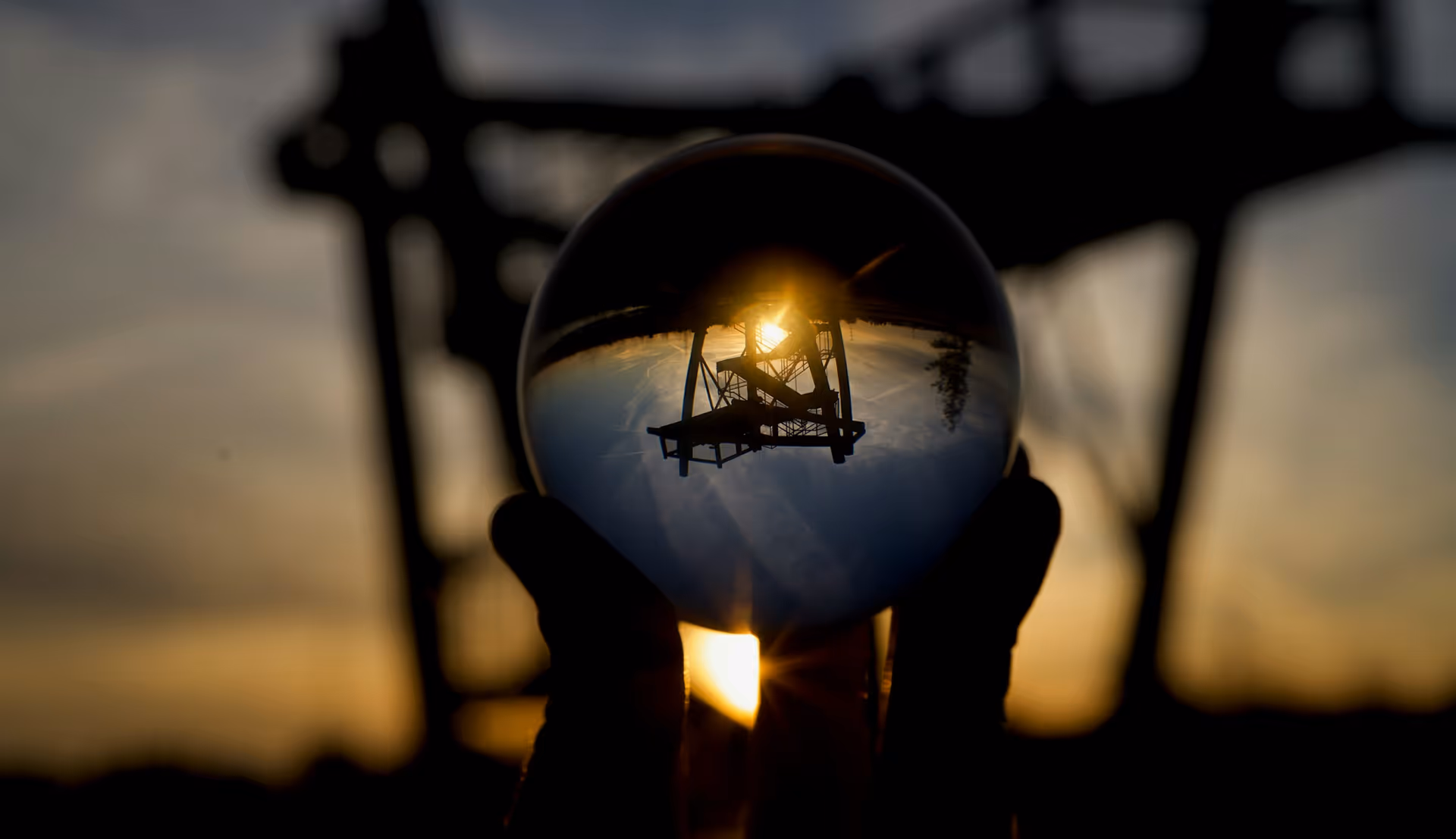 Silhouette of industrial structure reflected in a glass sphere held against a sunset sky.