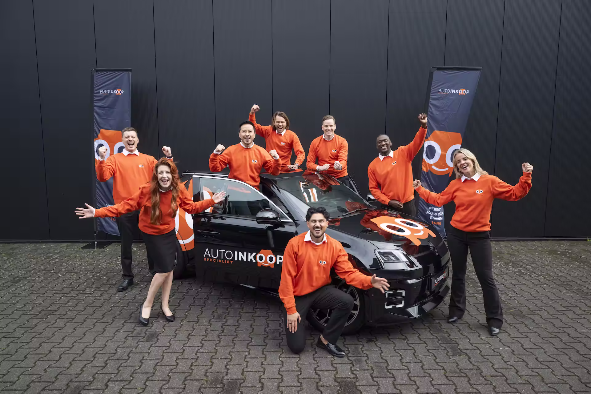 Team van zeven personeelsleden  in oranje truien poseert vrolijk rond een zwarte auto met een AutoInkoopSpecialist-logo.