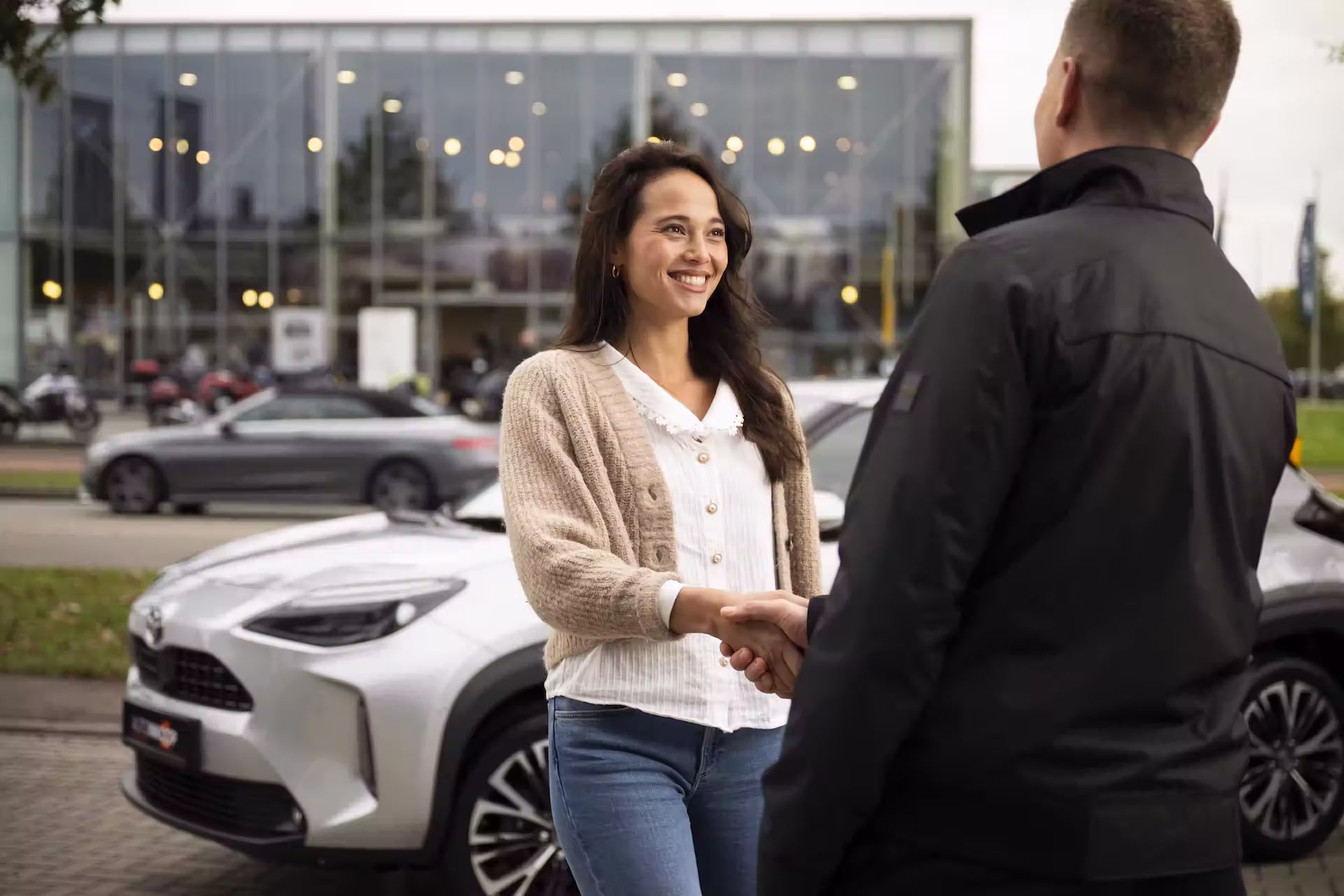 Vrouw minzaam lachend terwijl ze handen schudt met een man naast een zilveren auto buiten een dealer-gebouw.