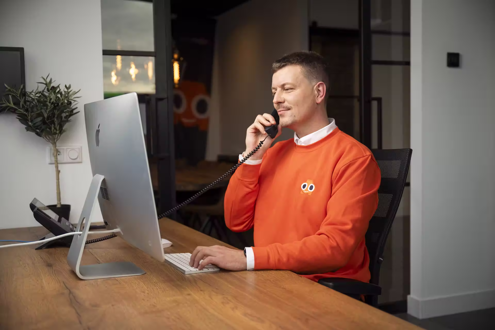 Personeelslid van AutoInkoopSpecialist met oranje trui die aan bureau zit en onderhandelt via de telefoon terwijl hij op toetsenbord typt.