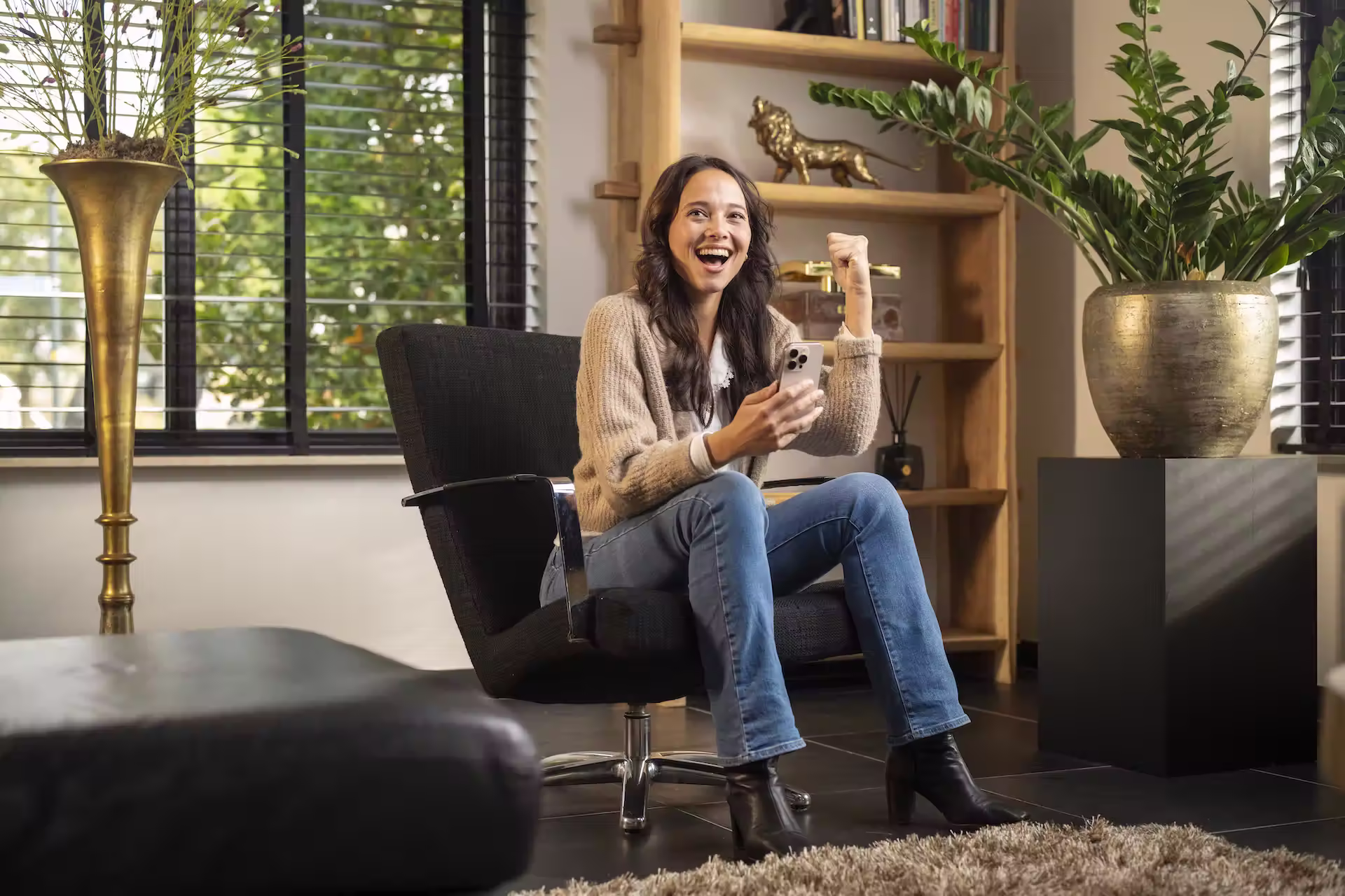 Vrouw zittend in een zwarte draaistoel, blij kijkend naar haar smartphone met een opgeheven vuist in een modern interieur.