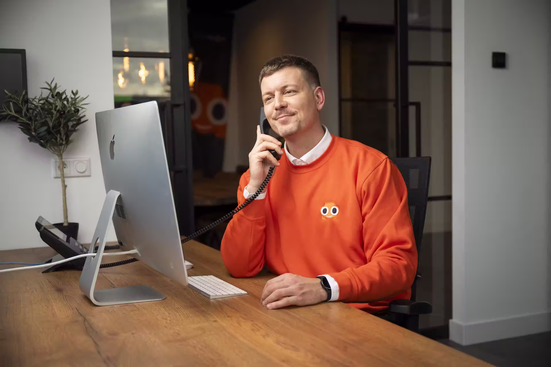 Personeelslid van AutoInkoopSpecialist met oranje trui die aan bureau zit en onderhandelt via de telefoon terwijl hij op toetsenbord typt.