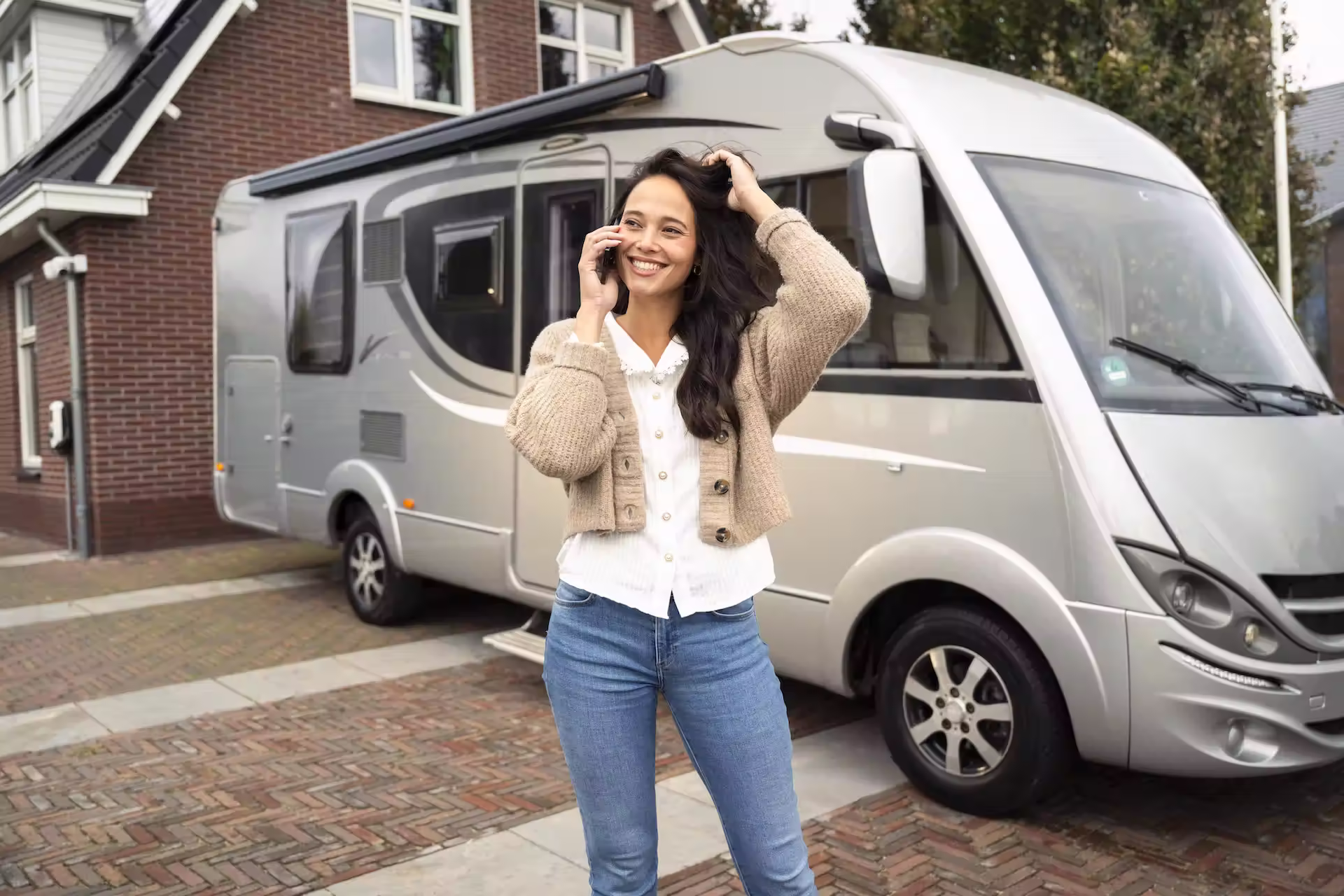 Vrouw die lacht en telefoneert terwijl ze staat voor een geparkeerde zilverkleurige camper bij een huis.