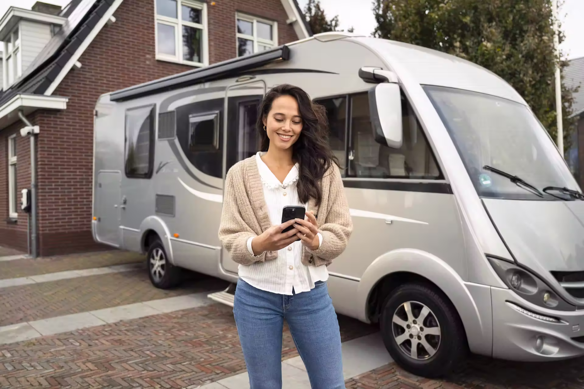 Vrouw met beige vest en spijkerbroek die lacht terwijl ze op haar telefoon kijkt naast een grijze camper geparkeerd voor een huis.
