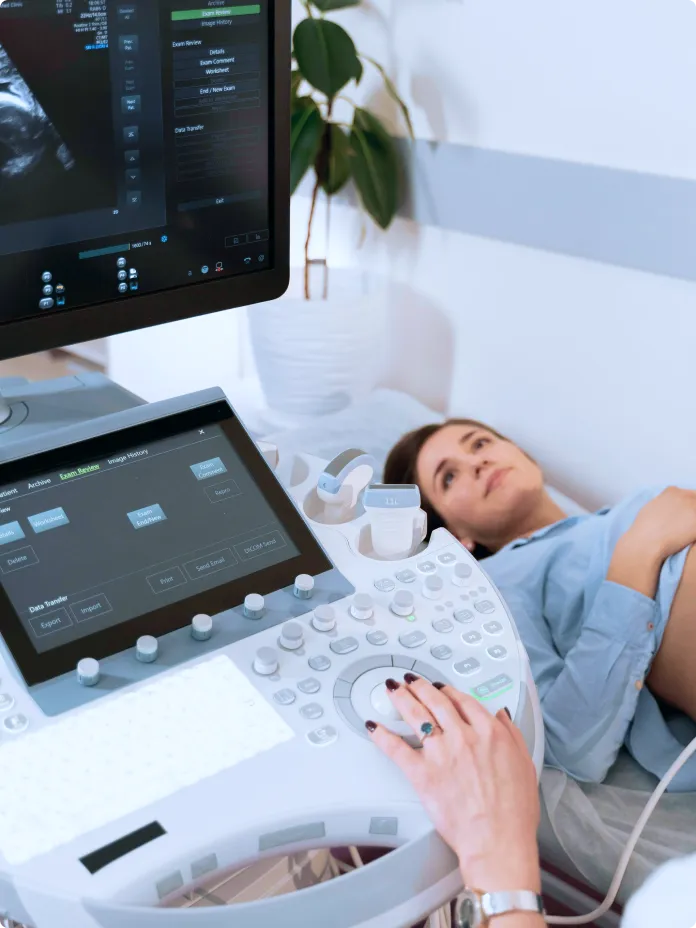 A pregnant woman lies on an examination table during an ultrasound scan. A medical professional operates the ultrasound machine, and a monitor displays the scan image. The woman is looking at the screen, smiling slightly, and resting her hand on her belly.