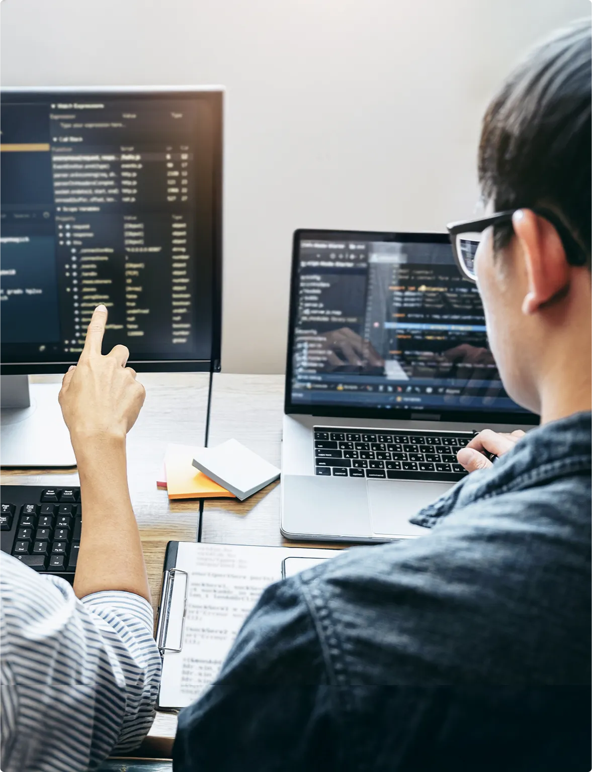 Two individuals collaborating at a computer, with a screen displaying lines of code in the background.
