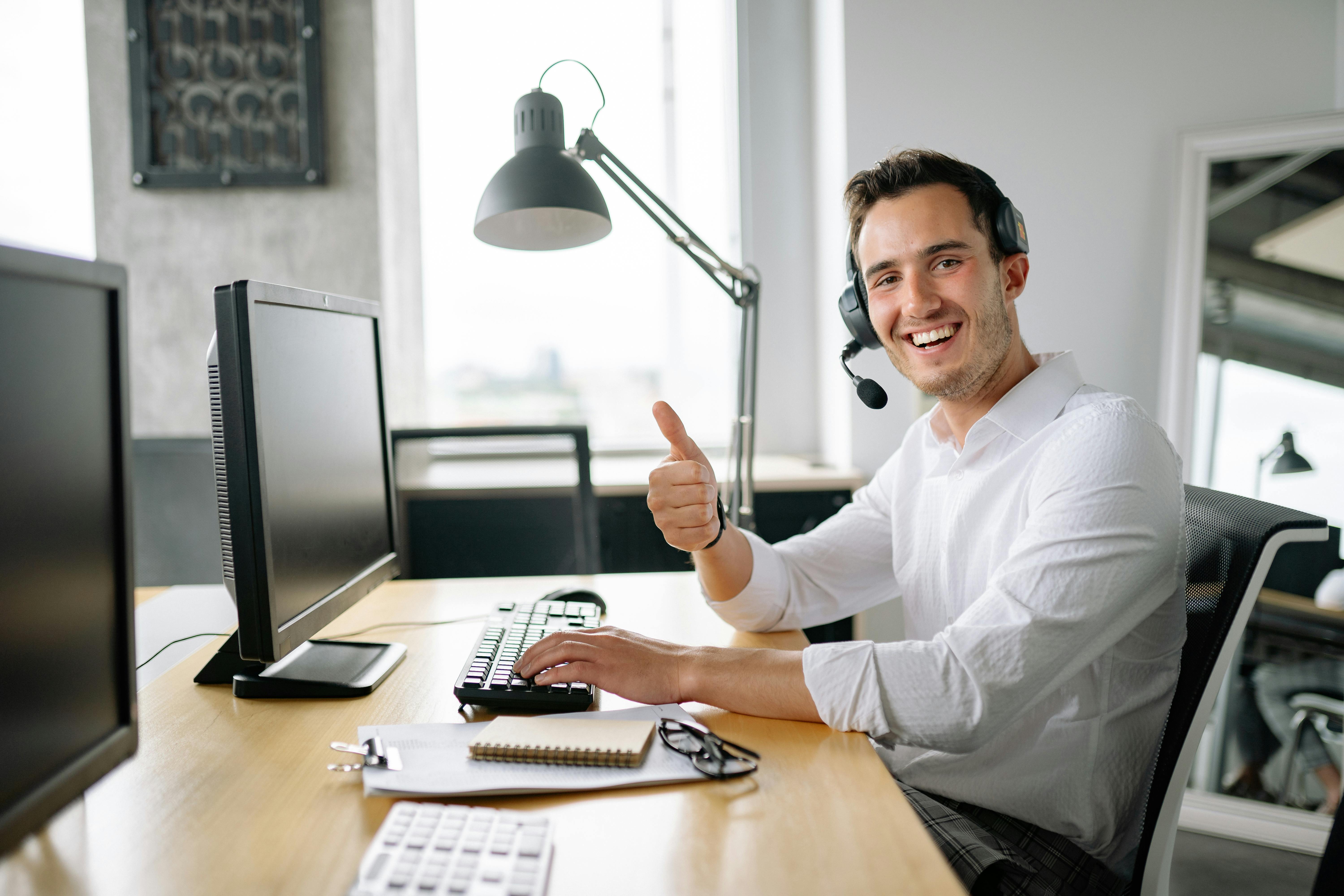 Lächelnder junger Versicherungsberater mit Headset sitzt an einem modernen Schreibtisch im Büro, zeigt Daumen hoch in die Kamera, spricht freundlich über ein Headset, vor ihm zwei Bildschirme, Notizblock und Tastatur – professionelles Umfeld für digitale K