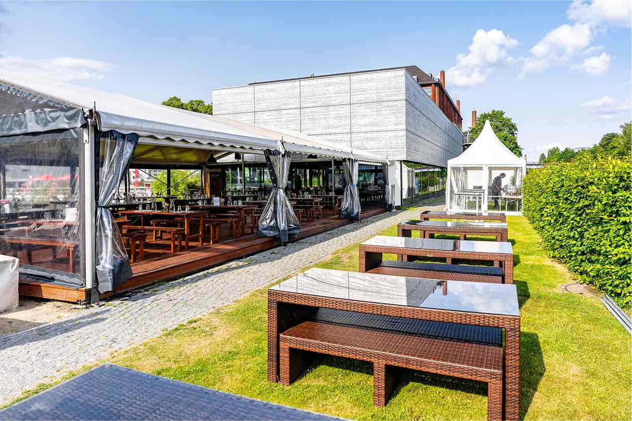 Pier 13 Berlin Tempelhof Eventlocation Außenbereich einer Veranstaltungslocation mit weißen Zelten, braunen Sitzgruppen auf Gras und einem modernen grauen Gebäude im Hintergrund.