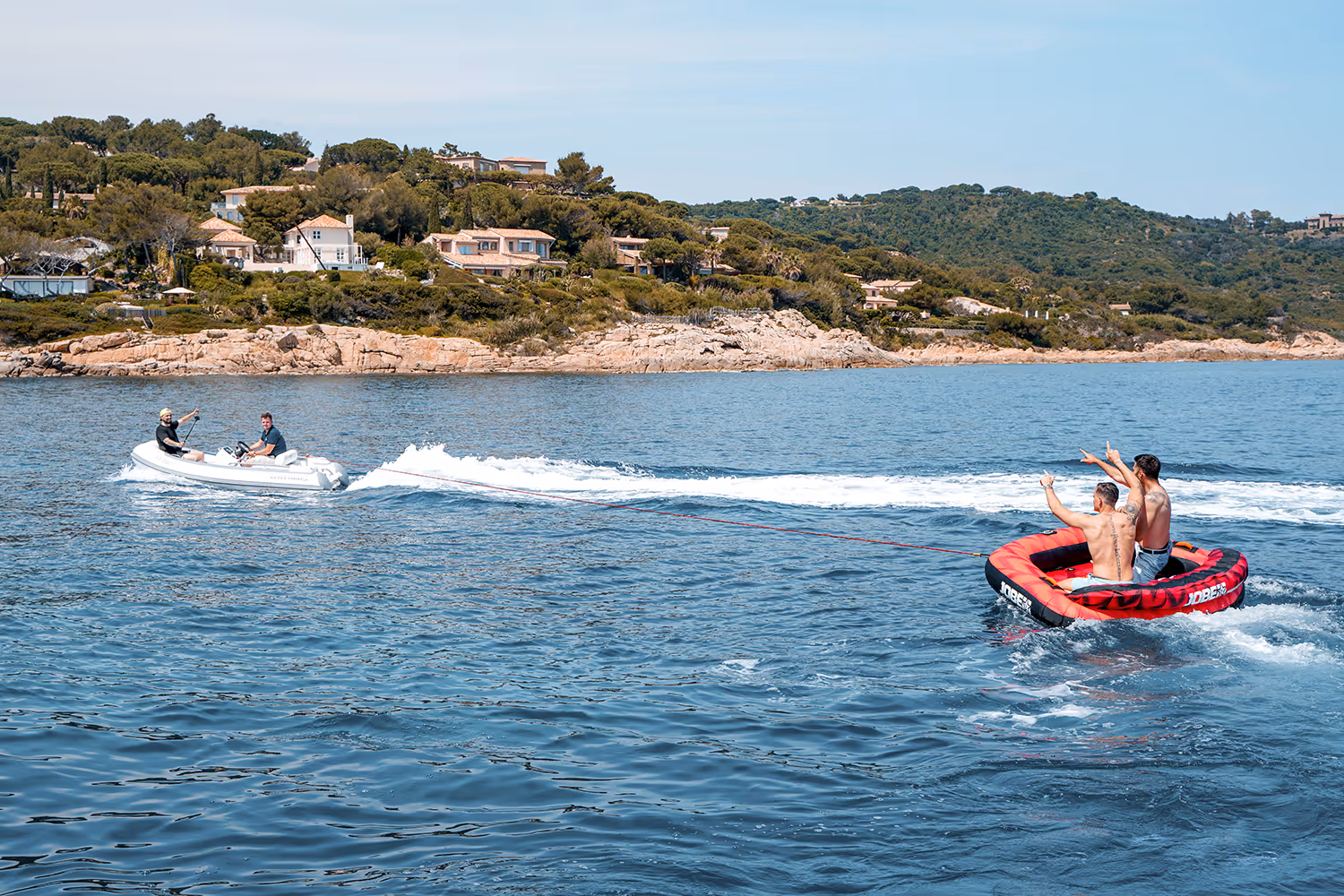 People riding a red inflatable towable tube behind a jet tender near a scenic coastline.