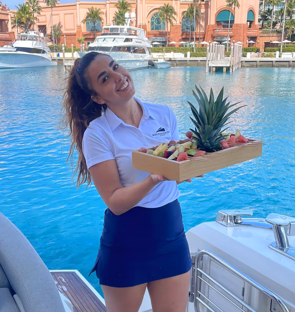 A young yacht stewardess in crew uniform is holding a wooden tablet with exotic fruit in her hands. She is on a yacht and the background shows Bahamas Paradise Island.