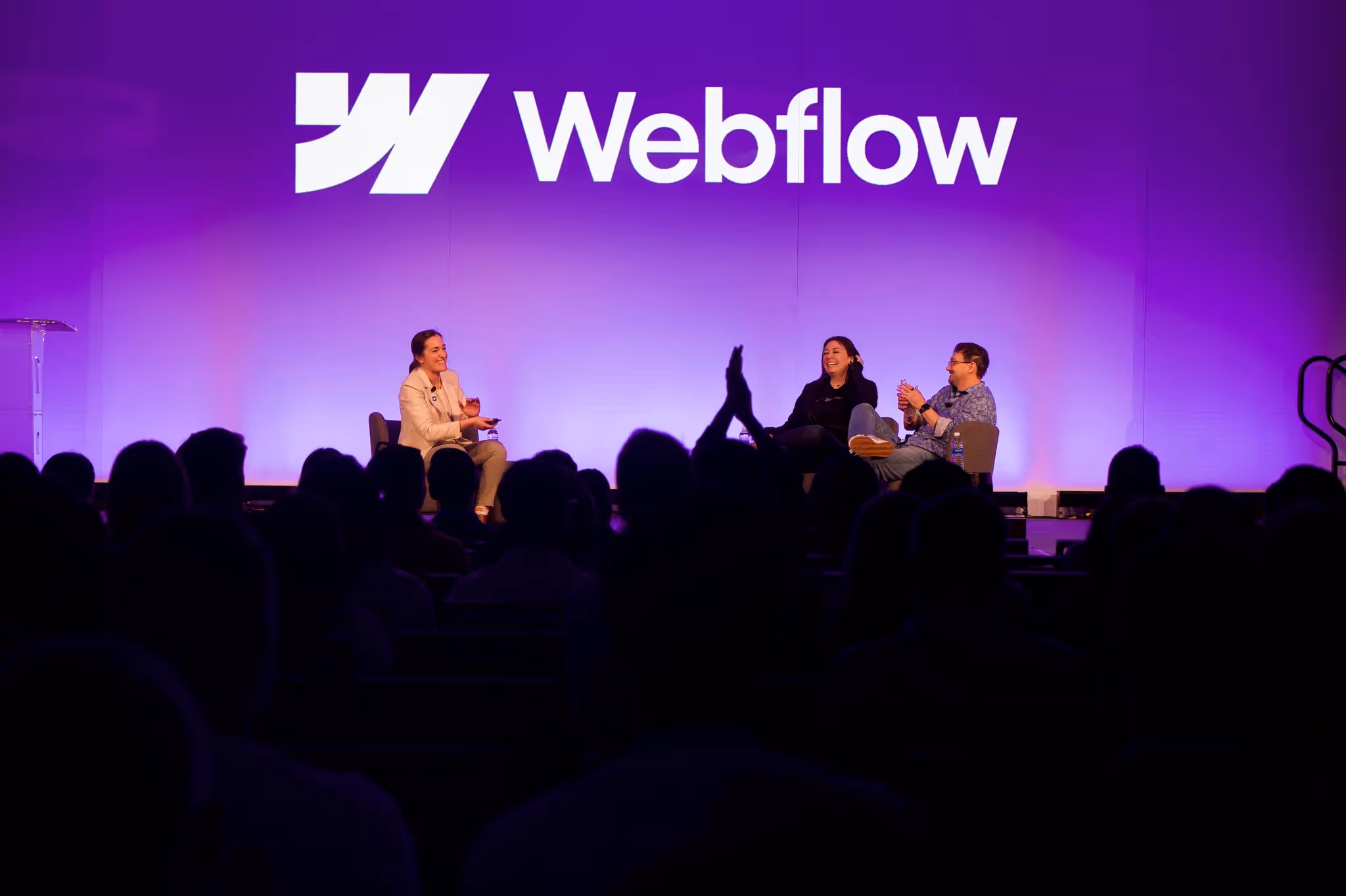 Three people seated on a stage having a panel discussion in front of an audience under a large Webflow logo on a purple-lit backdrop.
