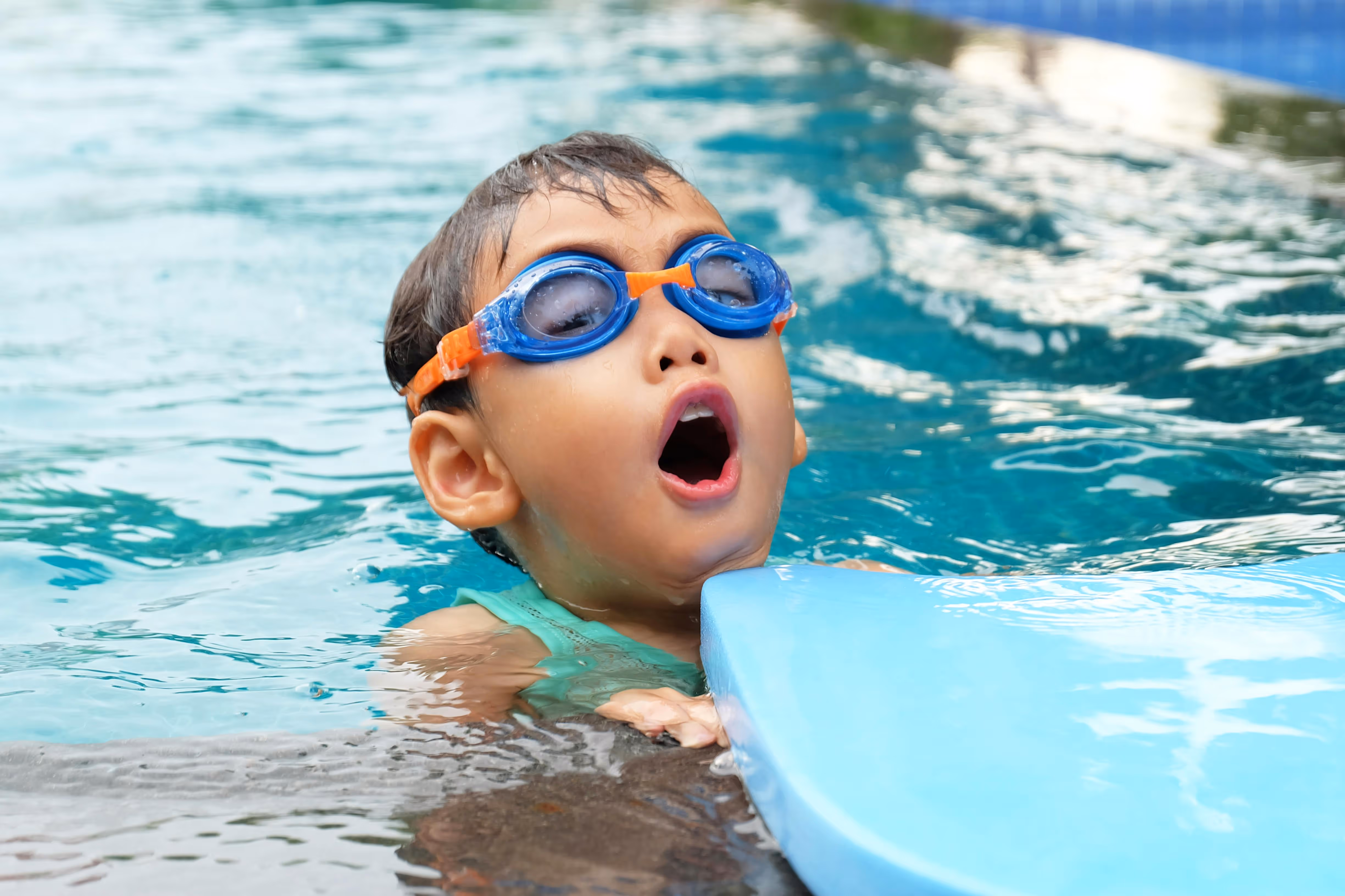 Toddler in goggles holding a kickboard in the pool.