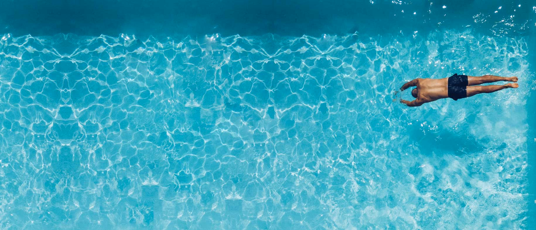 Man diving into clear blue swimming pool from above wearing black swim trunks.