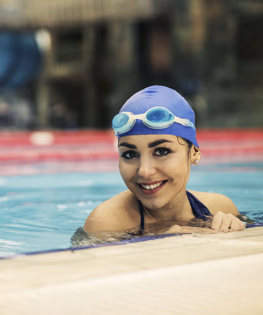 Woman in swim cap and goggles smiling at the pool edge.