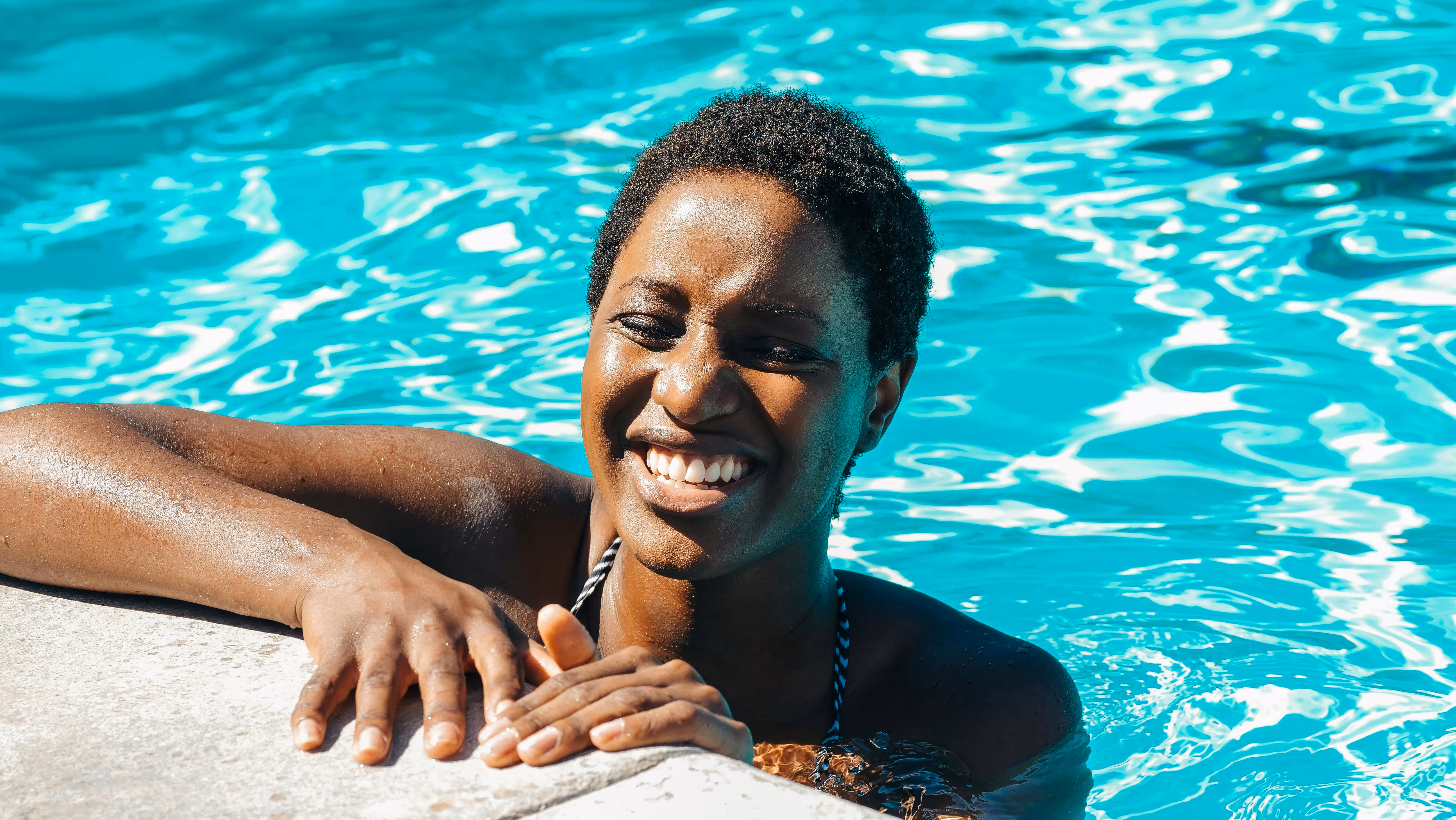 Smiling woman with short hair relaxing at the edge of a bright blue swimming pool.