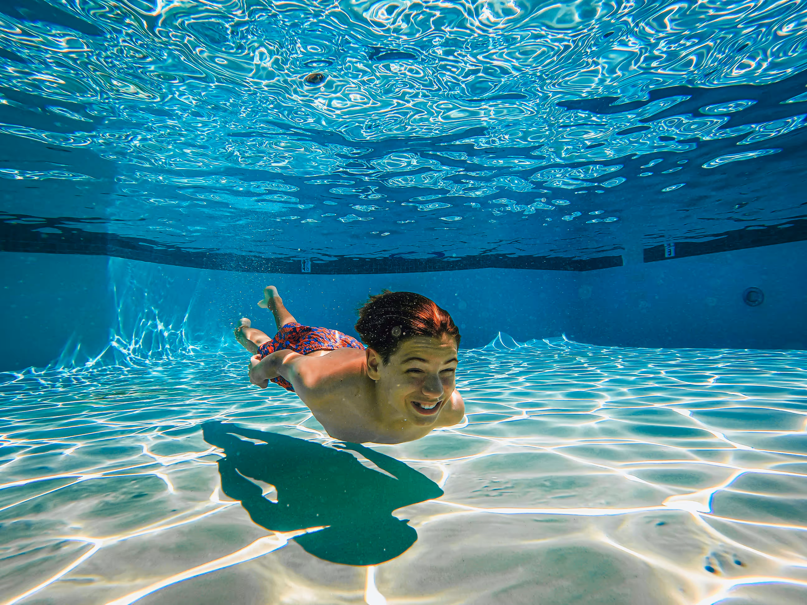 Boy swimming underwater in a clear pool with patterned sunlight on the floor.