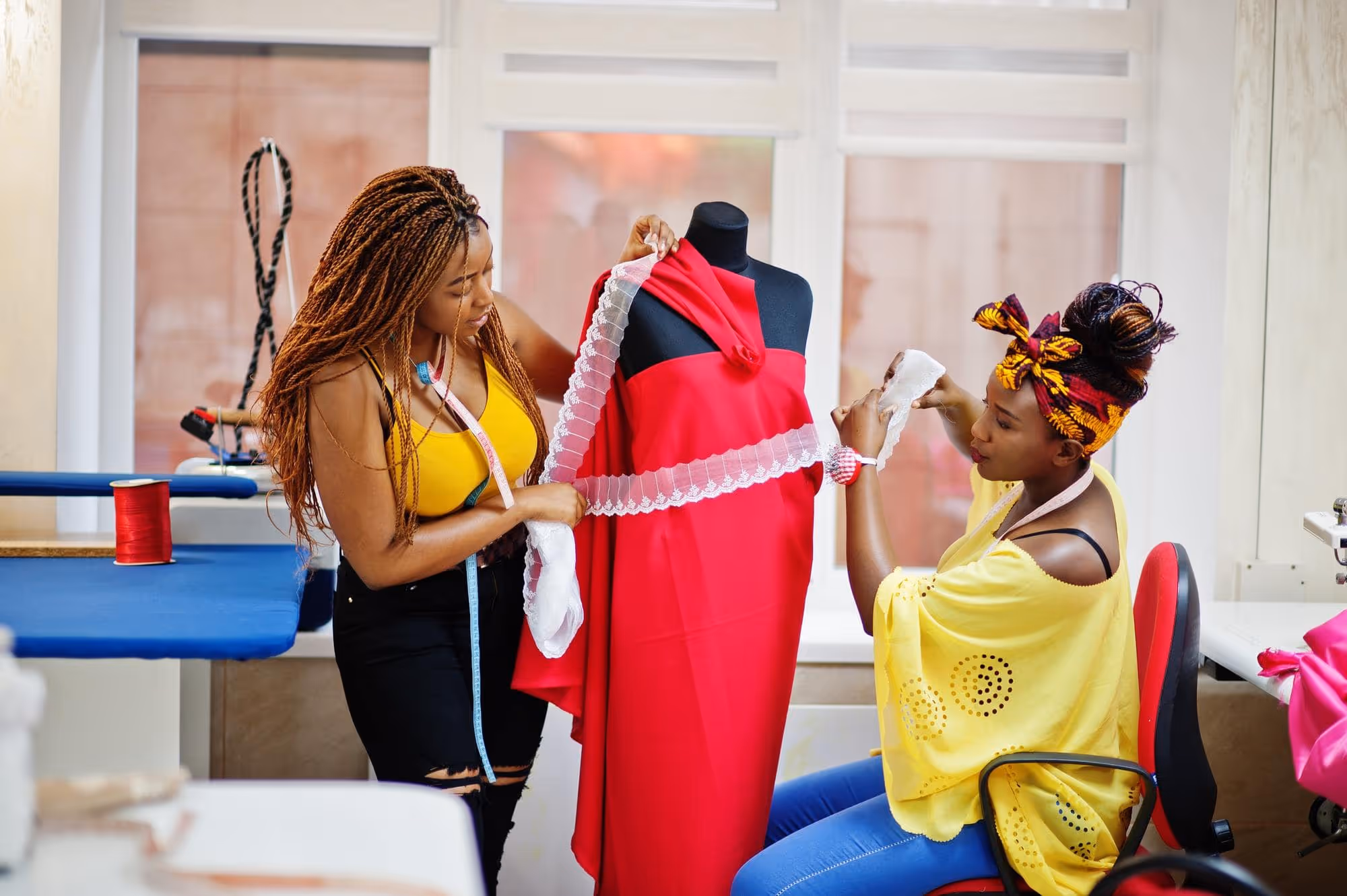 Two women working together measuring white lace fabric on a red dress displayed on a black mannequin.