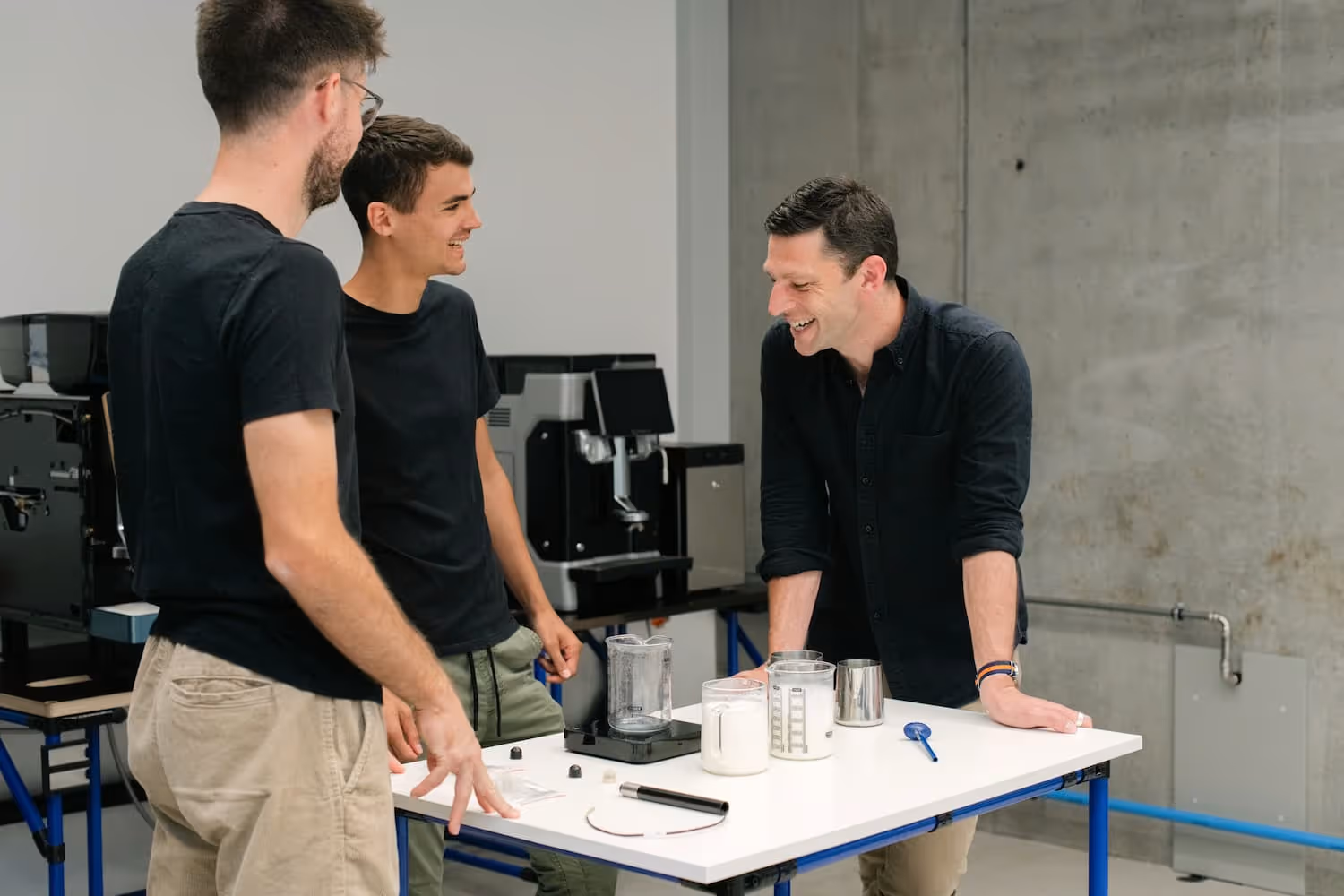Three men smiling around a table with milk jugs. 