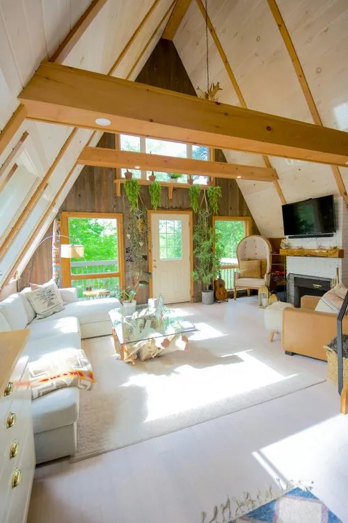 Bright, airy A-frame living room with large windows, greenery, cozy seating, and a natural wood coffee table.