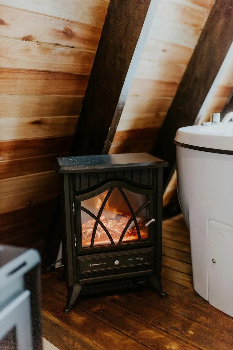 A cozy black electric fireplace in a wooden attic, glowing warmth beside a modern bathtub. Rustic wooden walls enhance the ambiance.